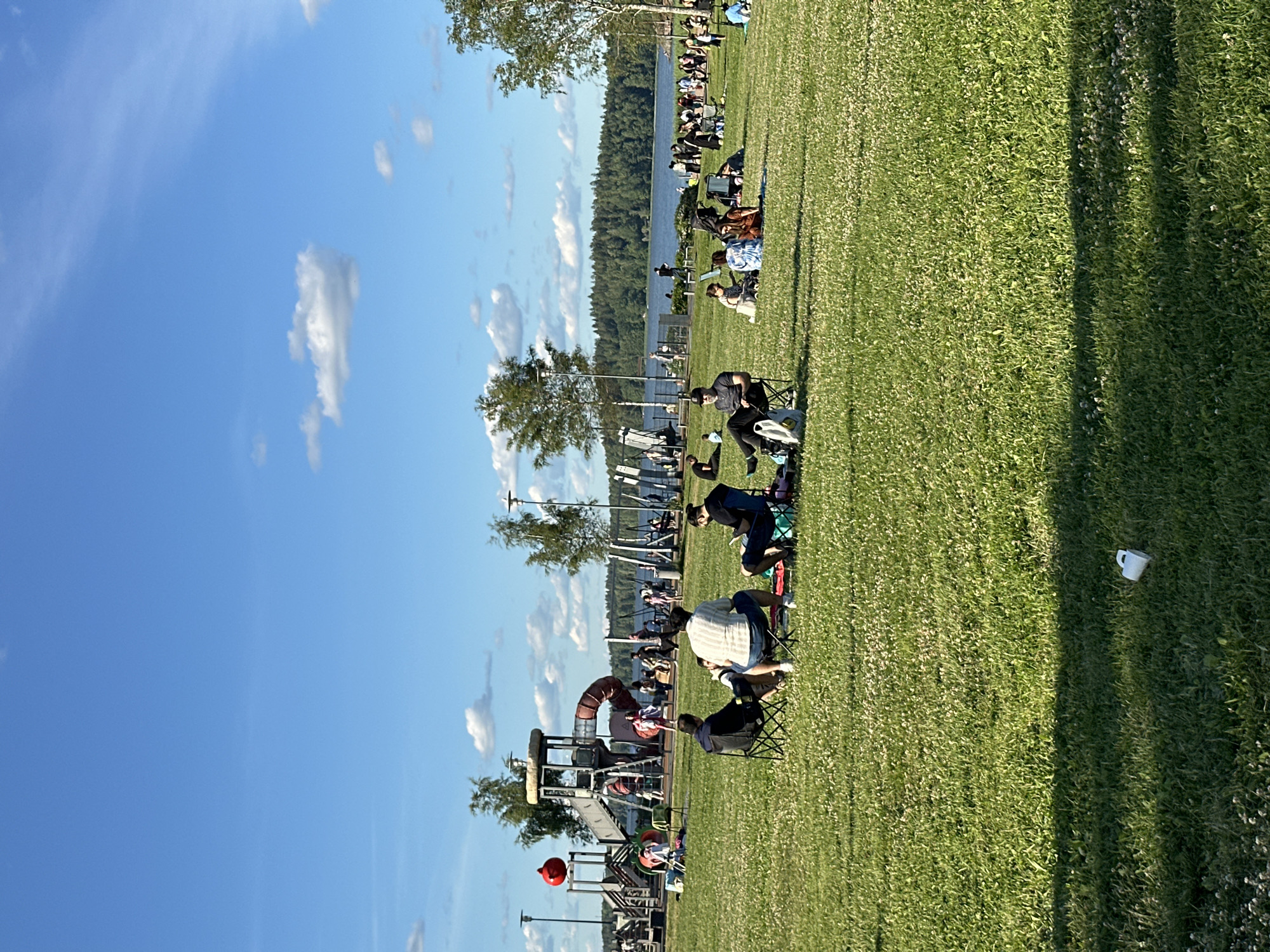 The image depicts a sunny day in a park with a body of water in the background. In the foreground, several people are scattered across a grassy area, some sitting in folding chairs and others on blankets. A playground structure with slides and climbing equipment is visible in the middle ground, with more people gathered around it. Trees line the edge of the water, and a distant treeline marks the horizon. The sky is blue with scattered white clouds. A white mug lies on the grass in the lower right corner of the image.