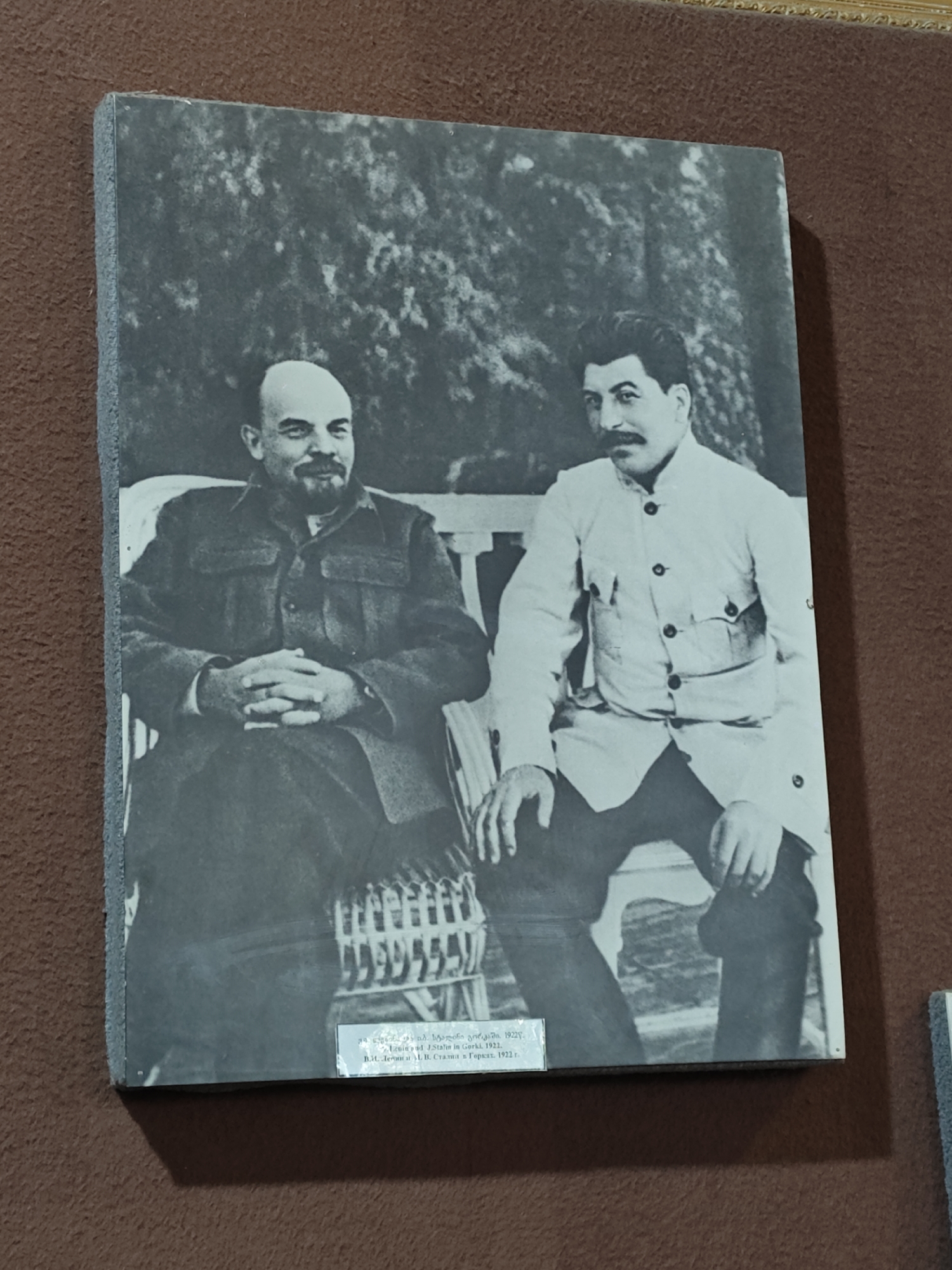 A black and white photograph depicts two prominent historical figures, Vladimir Lenin and Joseph Stalin, seated outdoors. On the left, Lenin, bald and bearded, wears a dark jacket and rests his clasped hands. On the right, Stalin, with dark hair and a mustache, is dressed in a light-colored, uniform-style jacket with multiple buttons and pockets. Both men are seated on simple benches, with Lenin on a wicker bench and Stalin on a wooden slat bench. The background features blurred foliage, suggesting a garden or park setting. Both subjects look towards the camera with neutral to slightly smiling expressions in this posed daytime shot. A small plaque at the bottom of the image contains text in Georgian, English, and Russian, stating "Lenin and J. Stalin in Gorki, 1922," identifying the subjects, location (Gorki), and the year the photograph was taken.