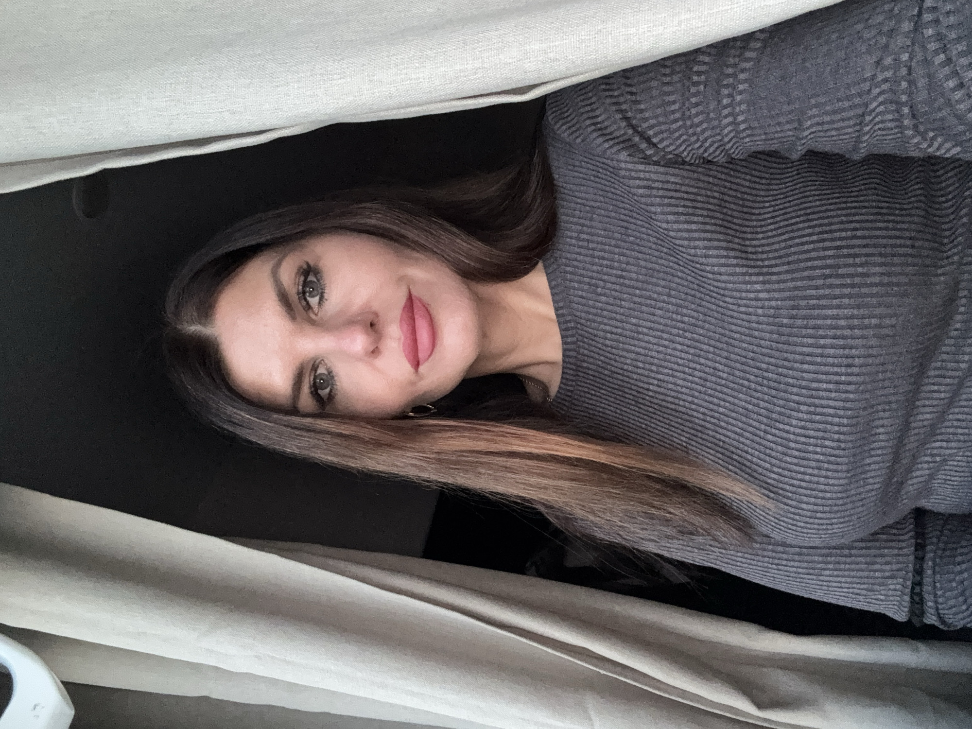 A woman with long brown hair and green eyes is looking at the camera, appearing from behind a light-colored curtain. She is wearing a grey ribbed sweater. The background behind the curtain is dark. There is no information in the image to identify the location as Chaikovsky, Russia.