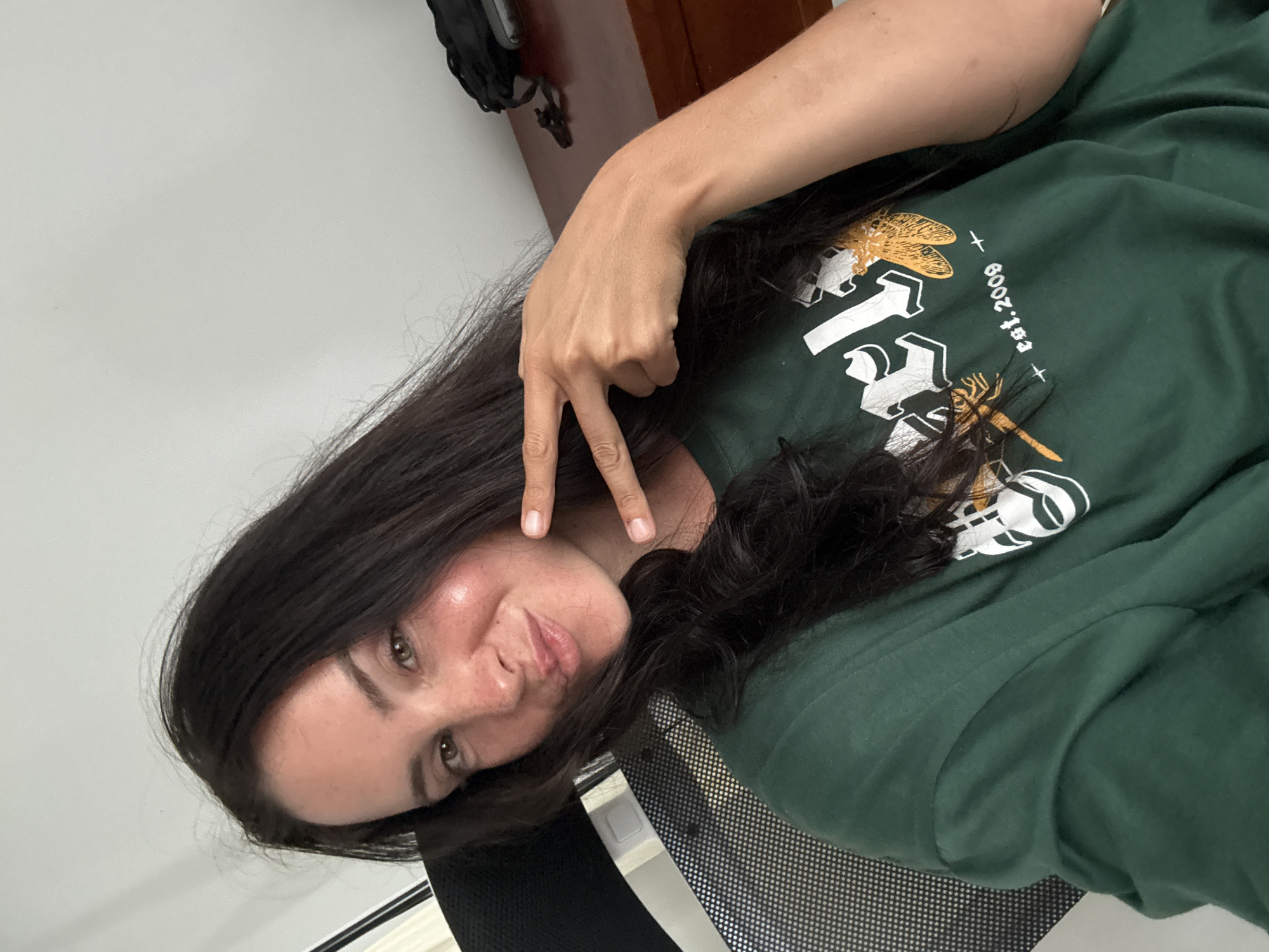 In a self-portrait taken indoors, a woman with dark hair and light skin is seated in an office chair. She is wearing a dark green t-shirt with a white graphic and the text "est. 2009" visible. Her right hand is raised, forming a peace sign. The background includes a desk made of wood, a black backpack on the desk, and a white wall. The lighting suggests an indoor setting, possibly an office or home office in North Kuta, Indonesia.