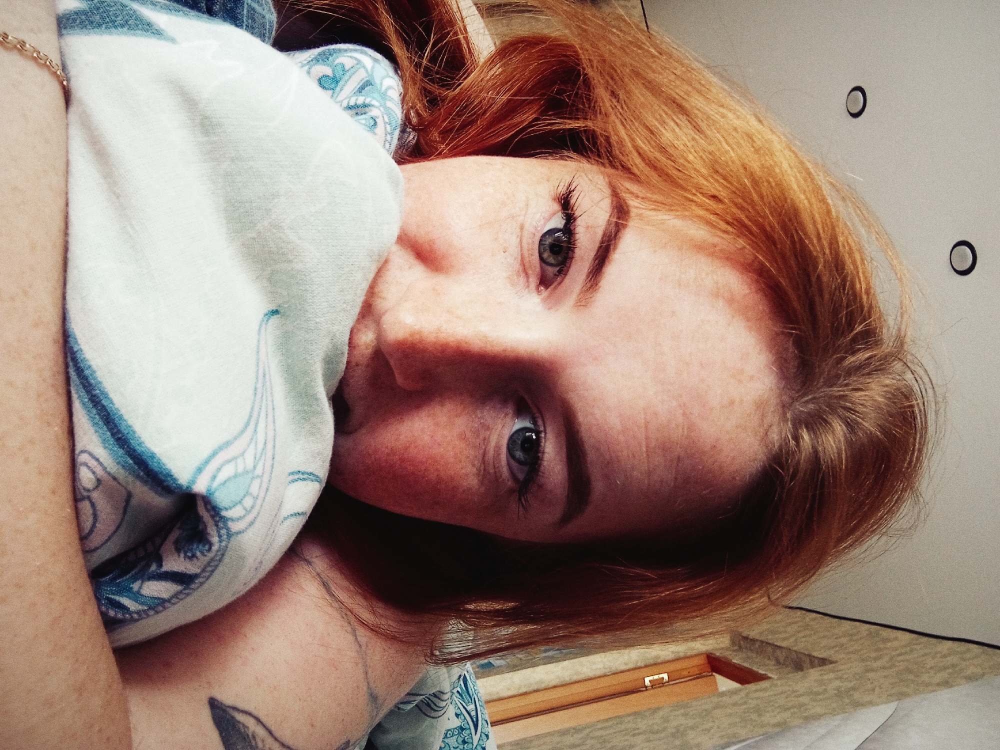 A close-up, slightly tilted, indoor photograph depicts a woman with red hair and fair skin. She has freckles and appears to be partially covered by a light-blue patterned fabric. A gold chain is visible on her arm. A tattoo is visible on her upper chest. The background shows a light-colored wall with two small, dark circles on it. A wooden structure is also partially visible in the background. There is no information to suggest this photo was taken in the city of Yurgamysh, Russia.