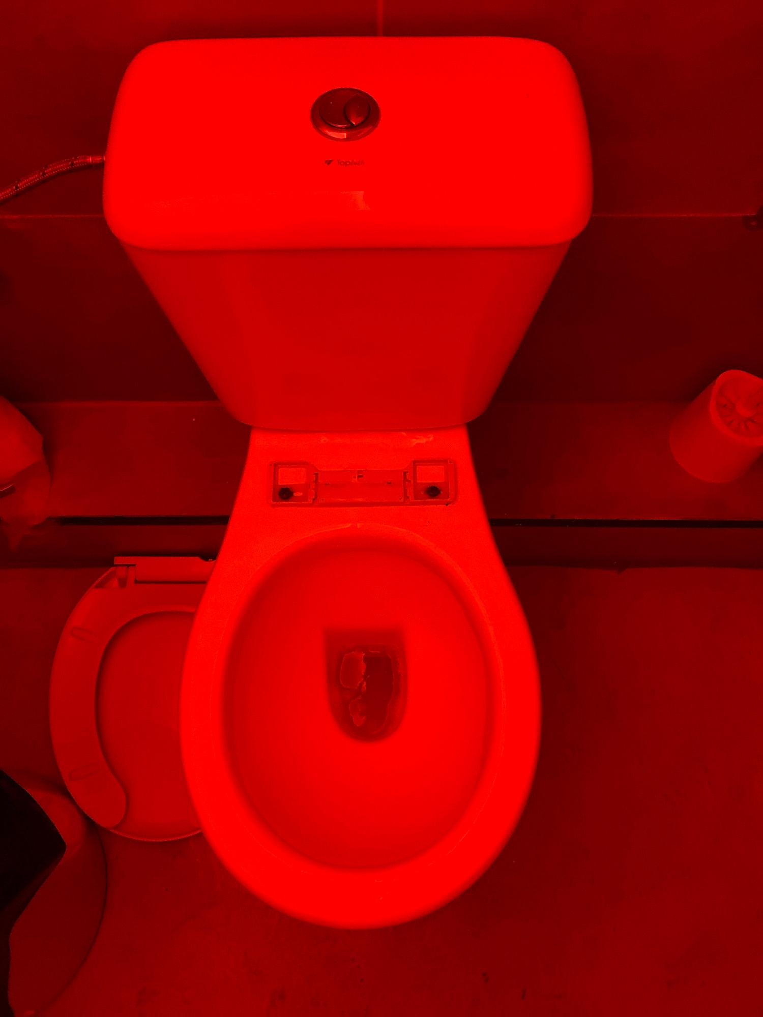 The image shows a red-tinted view of a toilet with its lid open and seat raised. The toilet is white ceramic and appears to be in a bathroom. There is a roll of toilet paper on a holder to the right of the toilet. A flexible water hose is visible on the left side of the toilet tank. The floor appears to be tiled or concrete. The overall scene is static and depicts a common bathroom fixture. The location is not discernible from the image.