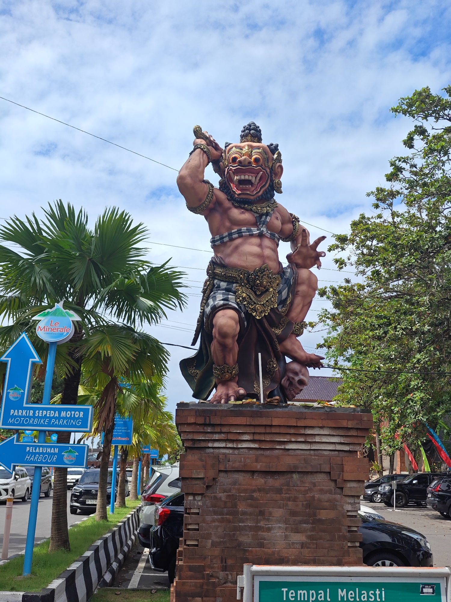 The image shows a large, ornate statue of a Balinese demon figure standing atop a brick pillar. The demon has a fearsome face, muscular build, and is adorned with traditional Balinese clothing and jewelry. It holds a weapon over its head and has a skull at its feet.
The statue is located on a roadside in Denpasar, Indonesia, indicated by the "Tempal Melasti" sign. Blue signs for "Le Minerale" are also visible, along with directional signs for "Parkir Sepeda Motor" (Motorbike Parking) and "Pelabuhan" (Harbour). Palm trees, other trees, and parked cars line the street. The sky is partly cloudy.