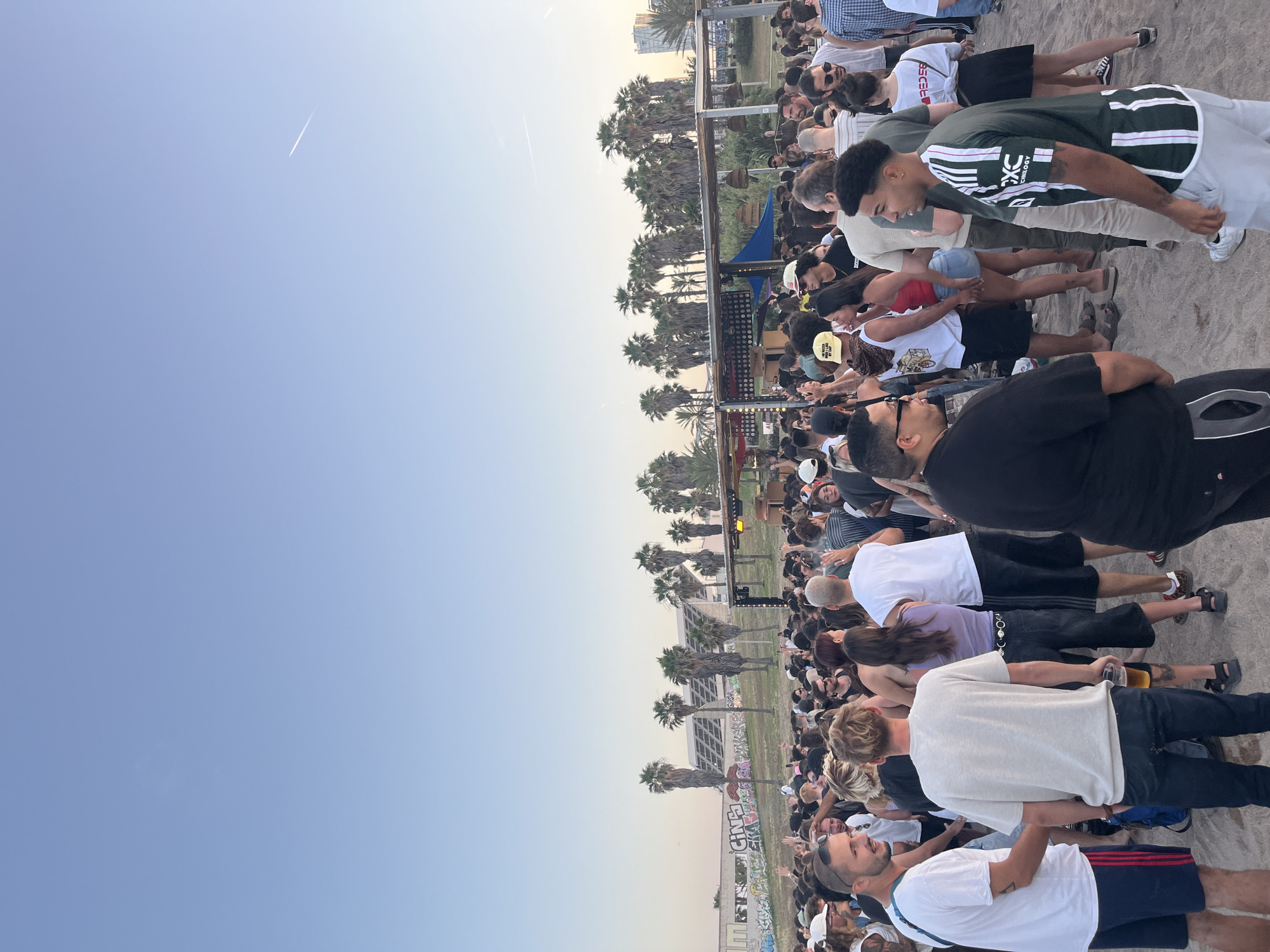 The image presents a large outdoor gathering of people on a sandy expanse in Sant Adrià de Besòs, Spain, likely a beach or park area adjacent to the Fòrum. The scene is illuminated by the clear, soft light of late afternoon or early evening, with a faint airplane contrail visible in the pale blue sky. In the mid-ground, a diverse crowd of individuals, dressed in casual summer clothing including t-shirts, shorts, and caps, are standing and interacting. Some are conversing, while others face towards a temporary stage or bar structure, identifiable by its canopy and visible lighting fixtures.
In the background, a line of palm trees stands between the crowd and a large, modern building equipped with a prominent array of solar panels. To the left of this structure, a wall covered in graffiti is visible. The ground is consistently sandy throughout the foreground and mid-ground.
Specific individuals in the foreground include a man on the far left wearing a white t-shirt and dark shorts with red stripes. To his right, another man, seen from the back, wears a light-colored t-shirt and dark pants, holding a clear plastic cup containing a yellow liquid, likely beer. On the right side of the foreground, a man wearing a green and white striped soccer jersey with "CXC technology" visible on the sleeve stands in profile. Further into the crowd, a woman in a red top and denim shorts is visible. Many people in the crowd are relaxed, with some appearing to be engaged in conversation or enjoying the event.