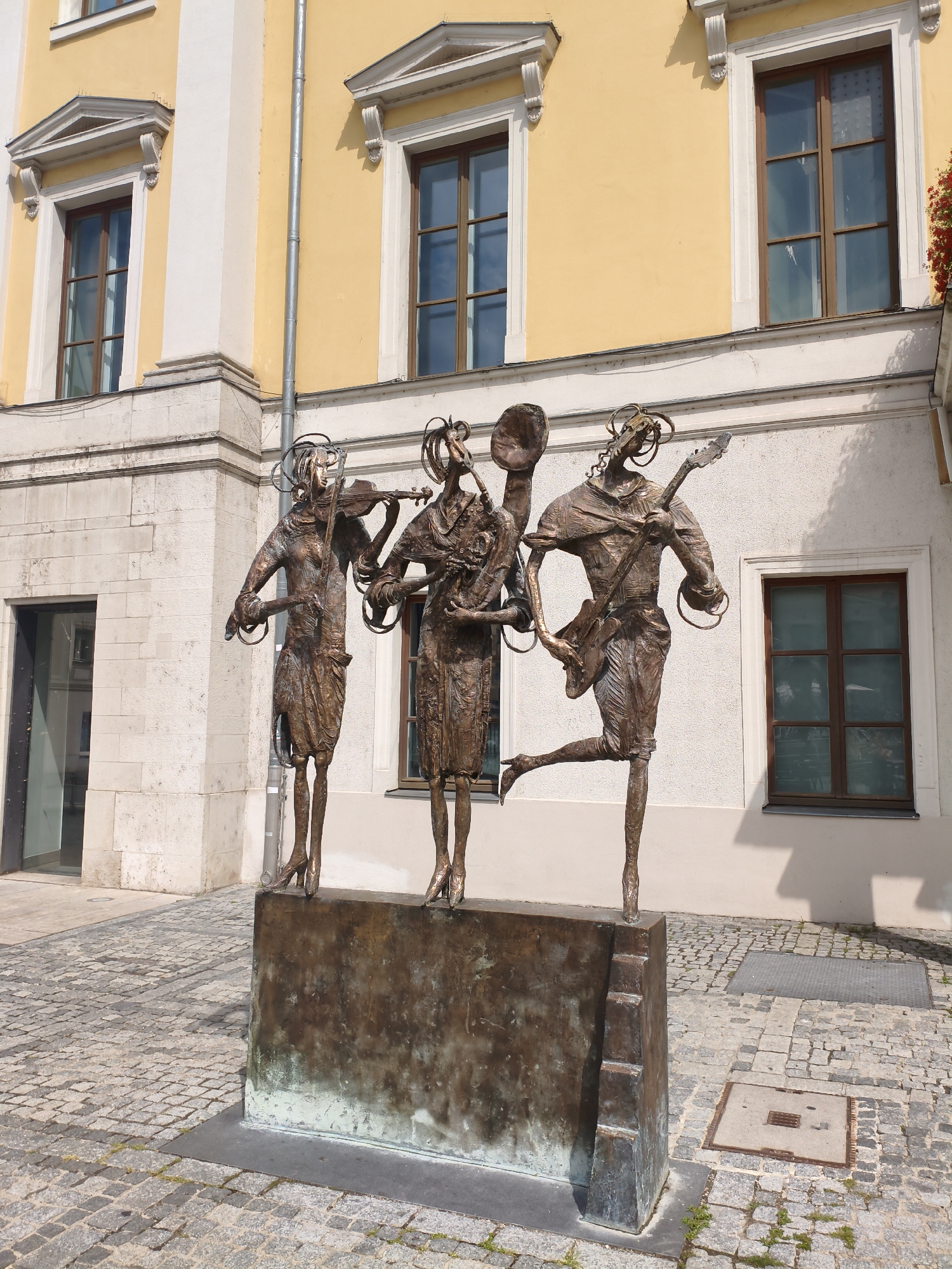 A bronze sculpture depicting three figures in the style of a band stands on a cobbled street in front of a yellow building. The figure on the left plays a violin, the middle figure holds a cymbal, and the figure on the right plays a guitar and has one leg raised. The figures have stylized wire-like elements on their heads. The building behind them features multiple windows with wooden frames and decorative architectural details. The ground is paved with dark grey cobblestones. This sculpture is located in front of the Thurn und Taxis Palais in Regensburg, Germany.