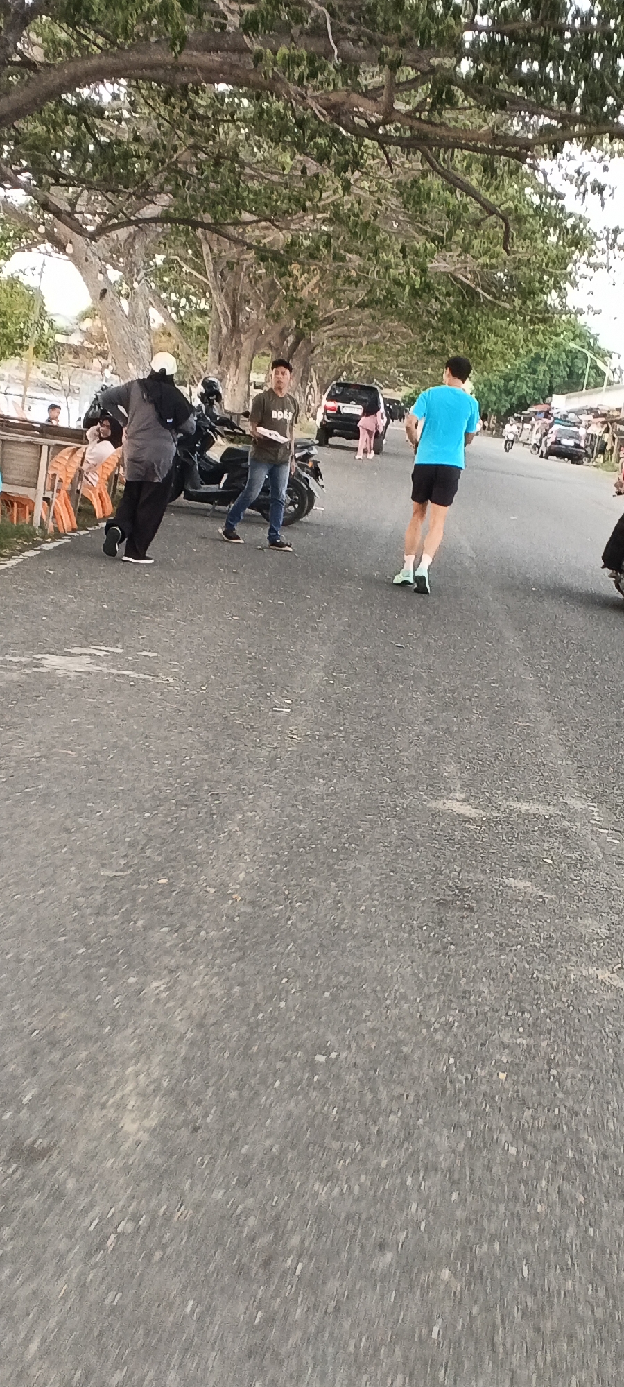 The image captures a street scene in Lhokseumawe, Indonesia, likely in the afternoon given the lighting. A man in a blue t-shirt and black shorts is skateboarding or rollerblading on the asphalt road, facing away from the camera. To his left, a man in a dark olive green t-shirt and blue jeans stands with a white rectangular object in his hands, looking towards the skateboarder. Behind him, a person in a grey outfit and white head covering walks away from the camera, leaning slightly, with a black bag slung over their shoulder. A dark-colored motorcycle is parked to the left of the man in the green t-shirt, with another motorcycle partially visible behind it. In the background, several cars are parked or driving on the road, including a black SUV. To the left of the road, there are orange plastic chairs and a wooden structure, suggesting a roadside seating area. Trees with dense green foliage overhang the scene from the left, casting shadows on the road. The overall environment appears to be an urban or semi-urban setting with a clear road surface and elements of everyday life.