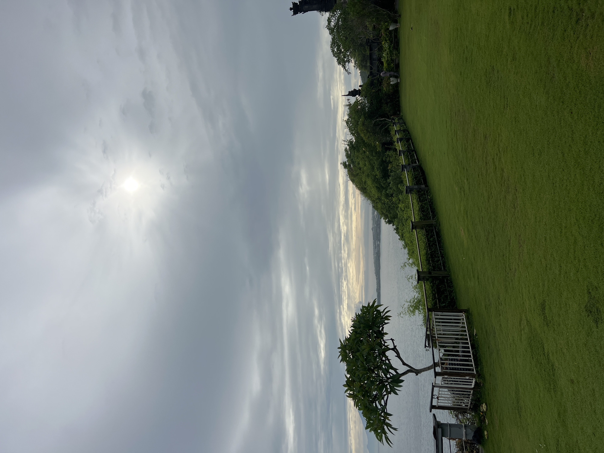 The image displays a serene coastal landscape, oriented horizontally, featuring a wide expanse of green grass, the sea, and a cloudy sky. The sun, a bright glare, is visible breaking through the clouds on the upper left side of the frame.
In the foreground, a meticulously maintained lawn of vibrant green grass occupies the right side of the image. A dark, wooden fence line runs along the edge of this grassy area, separating it from a dense band of trees and vegetation in the mid-ground. Beyond these trees, the calm waters of the sea stretch towards the horizon, reflecting the soft light of the sky.
On the far right, partially obscured by the trees, a tall, dark, and ornately detailed architectural structure, resembling a temple or traditional gate, stands prominently. Two indistinct figures are visible on the grass near this structure, facing towards the sea. In the lower left foreground, a small, leafy tree with dark green foliage grows near the water's edge, adjacent to a low, white railing or fence.
The scene suggests a peaceful daytime setting, possibly early morning or late afternoon, given the sun's position and the diffuse light filtering through the clouds. The weather is overcast or partly cloudy. There is no discernible activity taking place, beyond the presence of the figures. No visible text is present in the image. The architectural style and lush environment are consistent with the location context of Kuta Selatan, Indonesia.