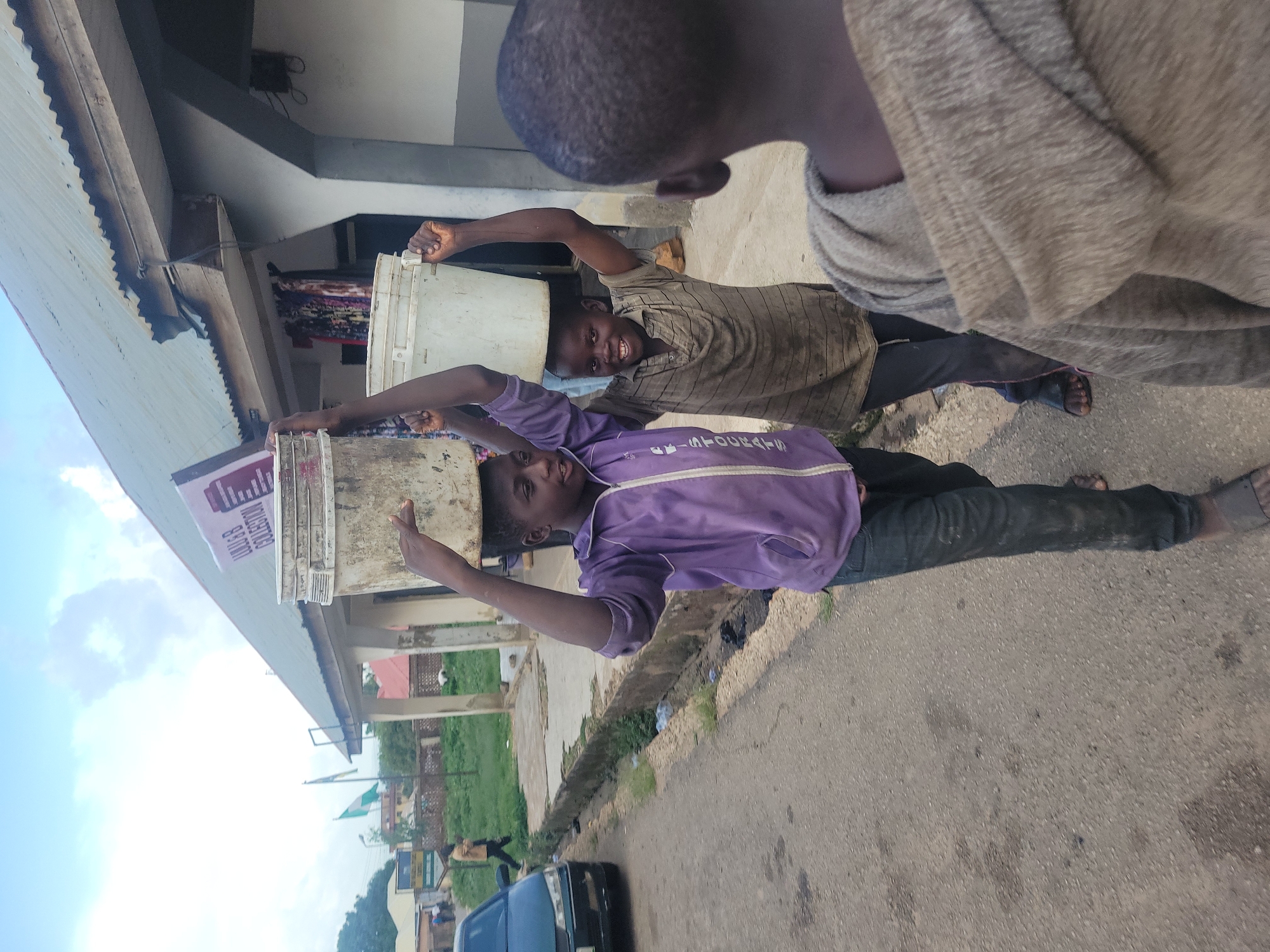 In Jos, Nigeria, two young boys are pictured carrying buckets of water on their heads. The boy on the left, wearing a purple jacket and dark pants, balances a large, dirty bucket. To his right, another boy in a striped shirt and dark pants smiles as he carries a white bucket. In the foreground, the back of a third boy in a hooded sweatshirt is visible. The boys are walking on a paved surface next to a dirt road lined with grass and debris. In the background, a building with a corrugated metal roof stands adjacent to a grassy area with trees and a distant sign. A dark blue car is parked on the left side of the road. The sky is overcast with hints of clouds.