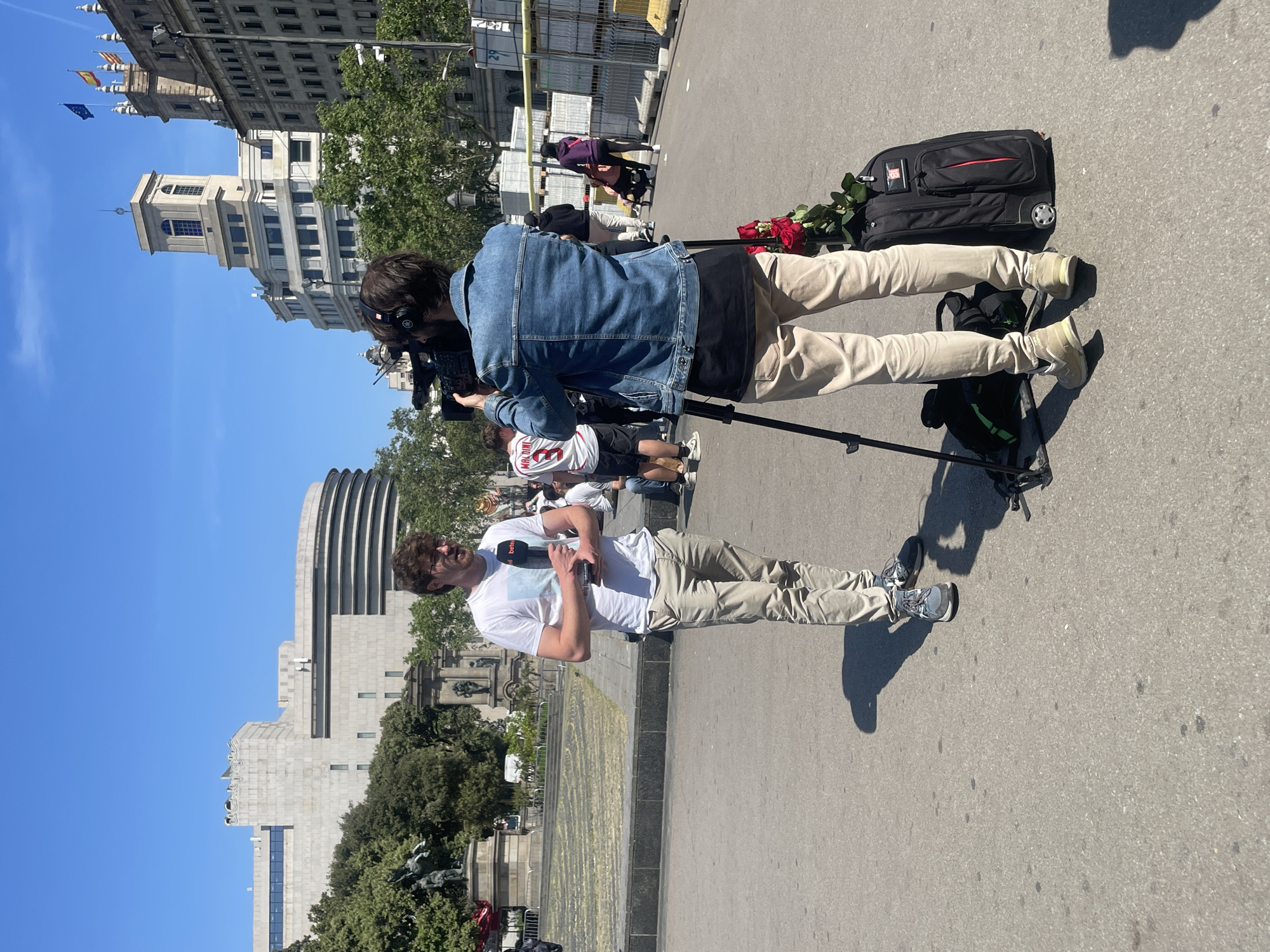 An outdoor video shoot is taking place on a paved street in Barcelona, Spain, under a clear, sunny sky. The scene features two men involved in the production, along with urban architecture in the background.
One man, seen mostly from behind, wears a denim jacket over a black t-shirt, beige pants, and headphones. He is operating a video camera mounted on a tripod, appearing to be the cameraperson. Next to him on the ground is a black, wheeled luggage bag with red accents, upon which a bouquet of red roses rests.
The second man, standing a short distance away, wears a white t-shirt, beige pants, and glasses. He holds a white microphone, which has the text "bwtv" visible on its black logo. He looks towards the right, suggesting he is presenting or reporting to an unseen audience or interviewer.
The background reveals a bustling urban environment with several notable buildings. These include an older, neoclassical-style building topped with spires, from which multiple flags are flying, including the Spanish, Catalan, and European Union flags. Another distinctive building with a modern, cylindrical tiered design is also visible. Trees and what appear to be public monuments or statues are scattered throughout the background. The bright sunlight casts distinct shadows on the ground.