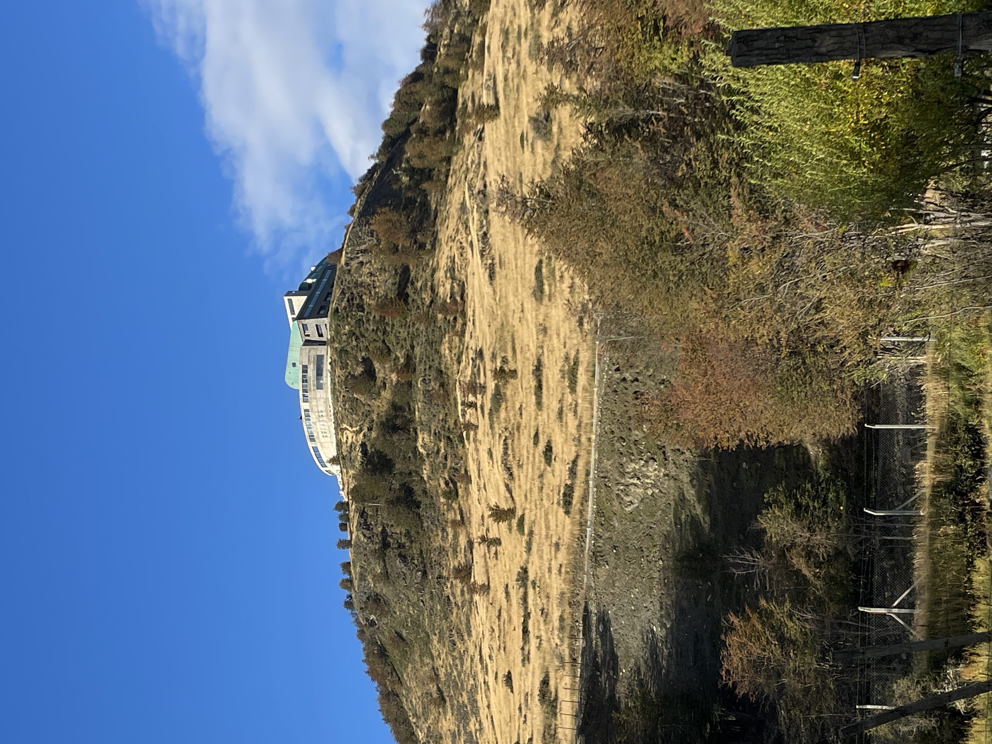 A medium, eye-level shot shows a hillside in Ushuaia, Argentina, against a clear blue sky with wispy white clouds at the top left. The hillside is covered in dry, yellowed grass and scattered shrubs and small trees, suggesting it is either late autumn or winter. At the top left, perched on the hill, is a multi-story building with a curved architectural design, appearing to be a hotel or apartment complex. A faint path is visible descending along the hillside, suggesting a walking or access route. In the foreground, a wooden fence runs parallel to the bottom of the frame, partially obscured by trees.