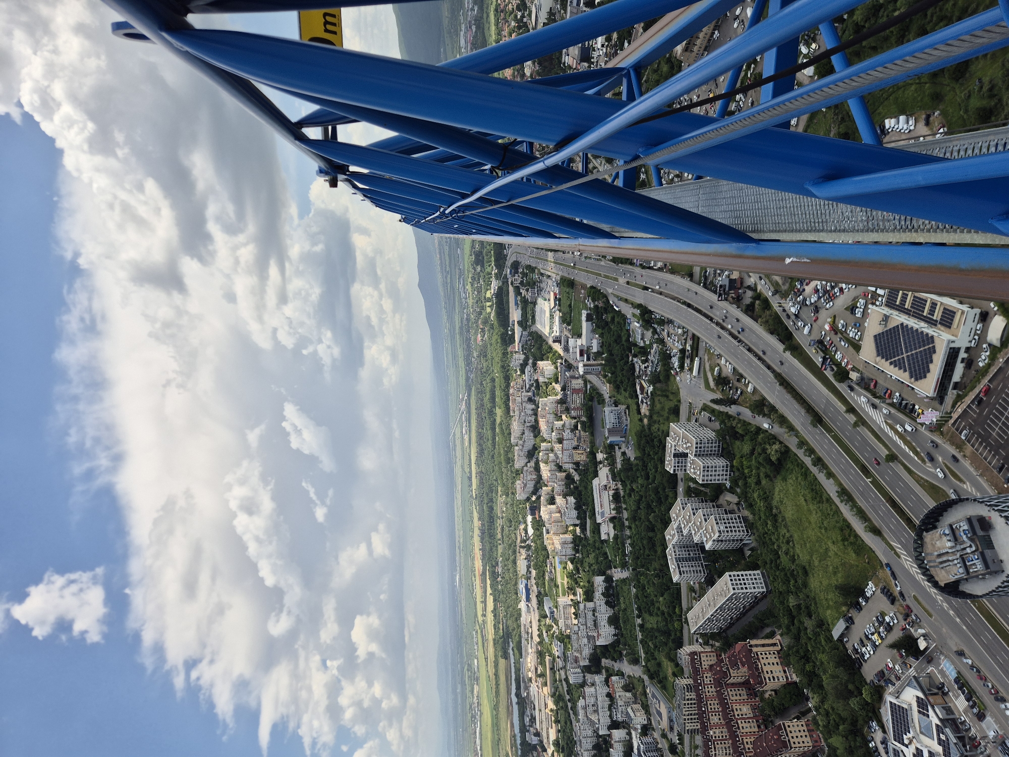 View from high altitude.
Blue steel construction lattice visible.
Clear sky and scattered clouds visible.
Landscape including a highway, city, and field visible below.
A building with solar panels on the roof is visible.
The scene shows Sofia, Bulgaria, with the Vitosha mountain range visible in the distance.