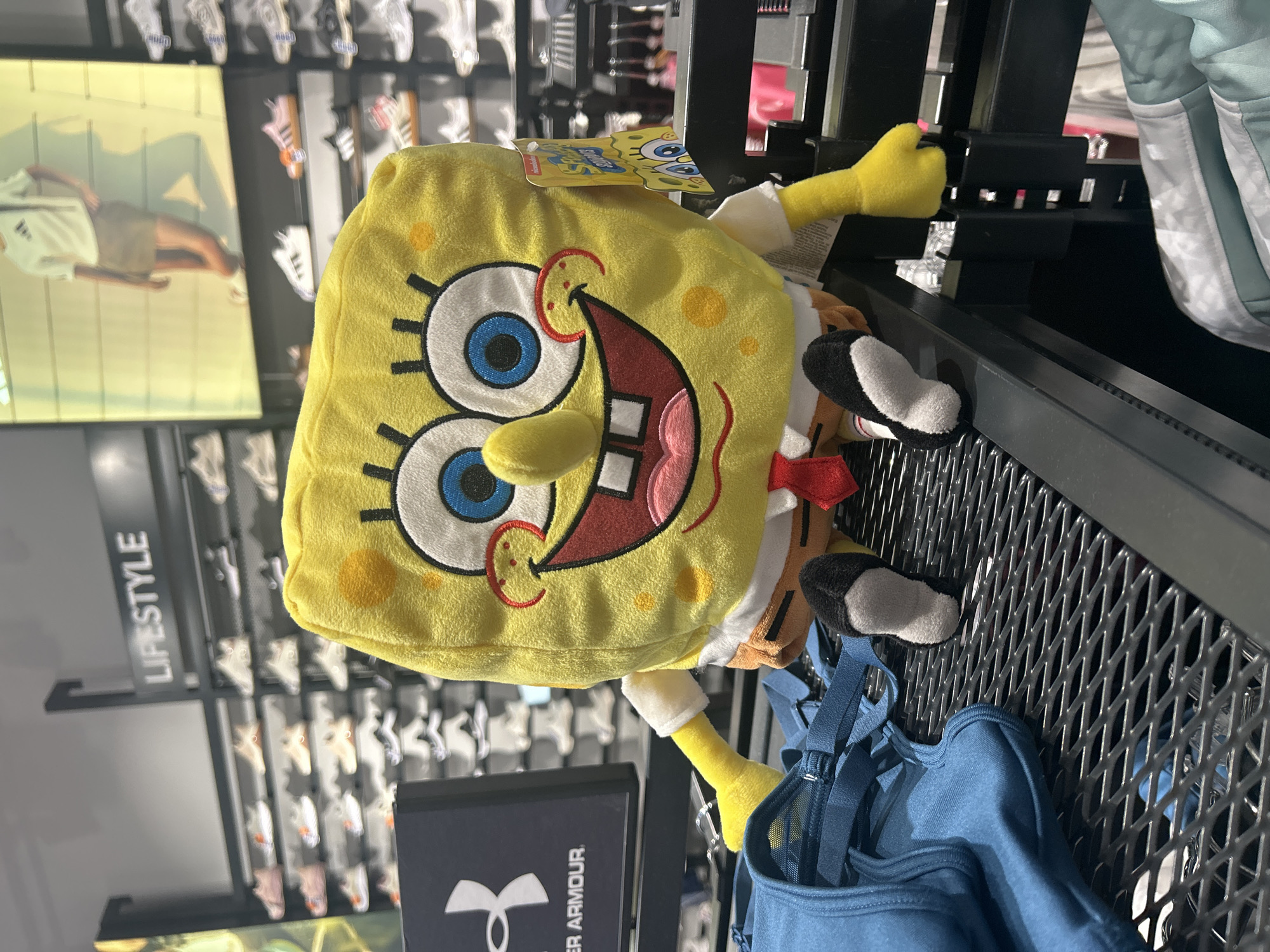 The image shows a Spongebob Squarepants plush toy lying on a metal display rack inside a store. The store appears to be a sporting goods retailer, as evidenced by the Under Armour logo on a nearby box and the display of athletic shoes in the background. The location of the store is unknown. The plush toy is yellow with a smiling face and is positioned next to a blue backpack.