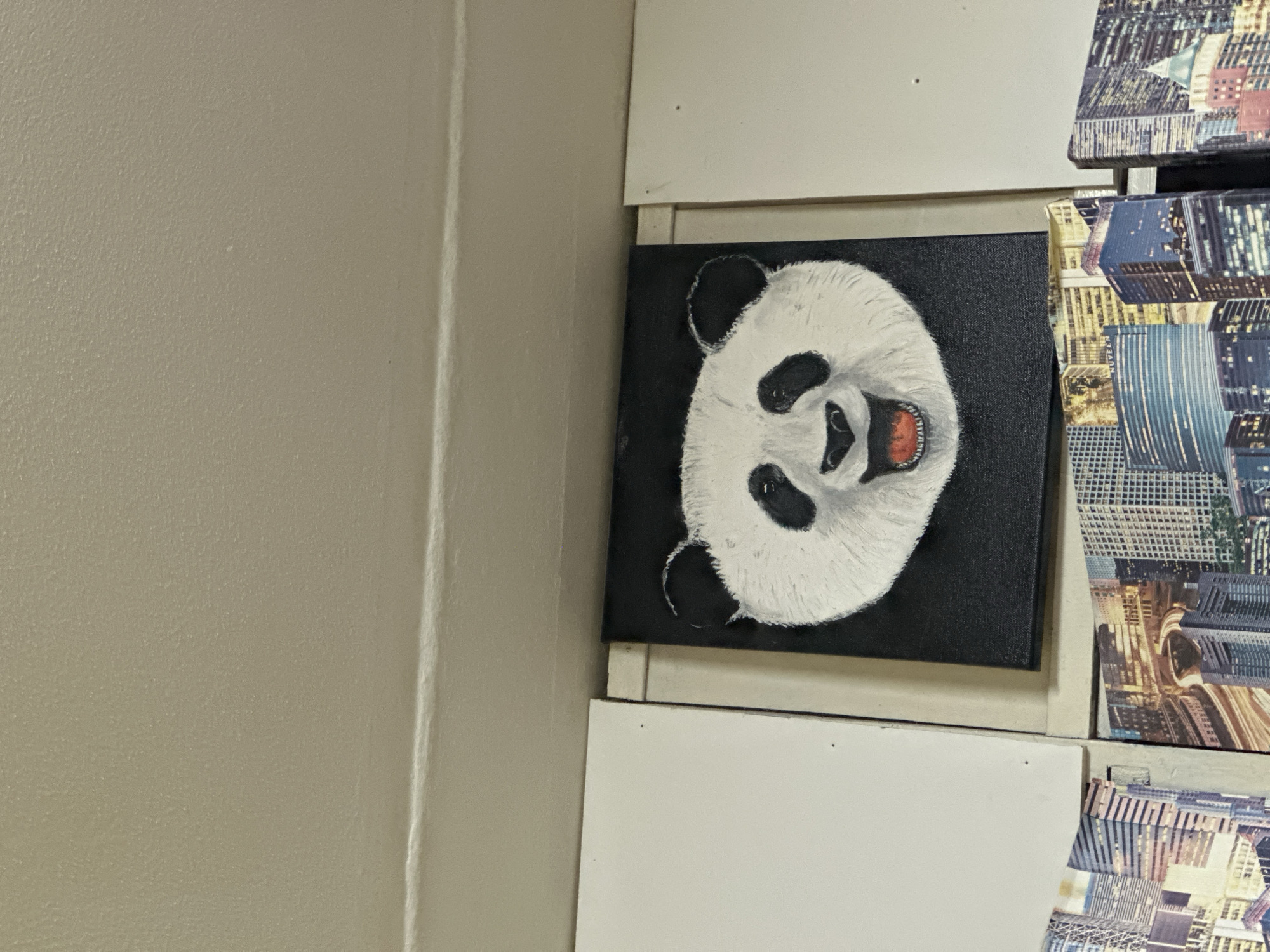 This image captures an indoor scene in Aktanysh, Russia, displaying multiple artworks mounted on a wall. The primary subject is a rectangular painting, centrally located in the lower half of the frame, depicting the head of a panda against a textured black background. The panda is rendered with white fur, black eye patches, ears, and nose, featuring an open mouth with visible red tongue and teeth, conveying a smiling or laughing expression.
To the immediate left and right of the panda painting are partially visible, plain white rectangular panels or canvases. Below the panda painting, and extending to the bottom edge of the frame, are sections of two different canvases displaying complex urban cityscapes, illuminated at night or dusk, with numerous buildings and visible light trails on roads. Text resembling "NUVEEN" is discernible on a building within the cityscape on the lower left.
The upper half of the image consists of a light beige or tan-colored wall or ceiling surface, which exhibits a fine, uniform texture. A horizontal seam or joint is visible across this upper section. The scene is evenly lit, with no individuals, explicit actions, or interactions present.