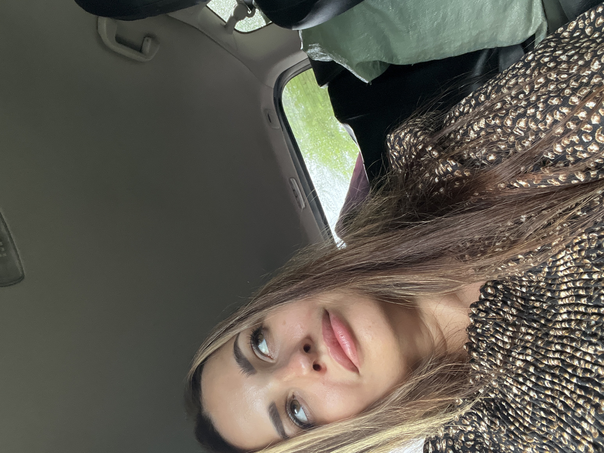 A young woman with long, dark blonde hair and makeup is captured from the chest up, looking to her left. She is inside a vehicle, as indicated by the car's ceiling and a handle visible in the upper portion of the frame. The woman wears a patterned top with a ruffled neckline. The background is blurred, showing a green, leafy landscape through the car window, suggesting an outdoor environment. A large, light green duffel bag is visible in the back seat, to the right of the woman. The overall lighting is natural and appears to be daytime.