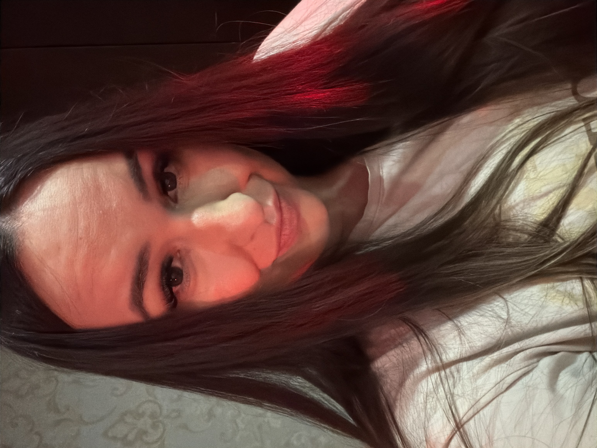 A young woman with long, dark hair is captured in a close-up, forward-facing selfie. She is wearing a light-colored t-shirt and her face is illuminated by a red light source, casting a red hue across her forehead, cheeks, and hair. Her eyes are open, and she has long, dark eyelashes. Her lips are slightly parted in a subtle smile. The background appears to be an indoor setting with textured wallpaper on the left and a dark surface on the right. There are no other people or prominent objects in the frame. The image is taken from a slightly upward angle.