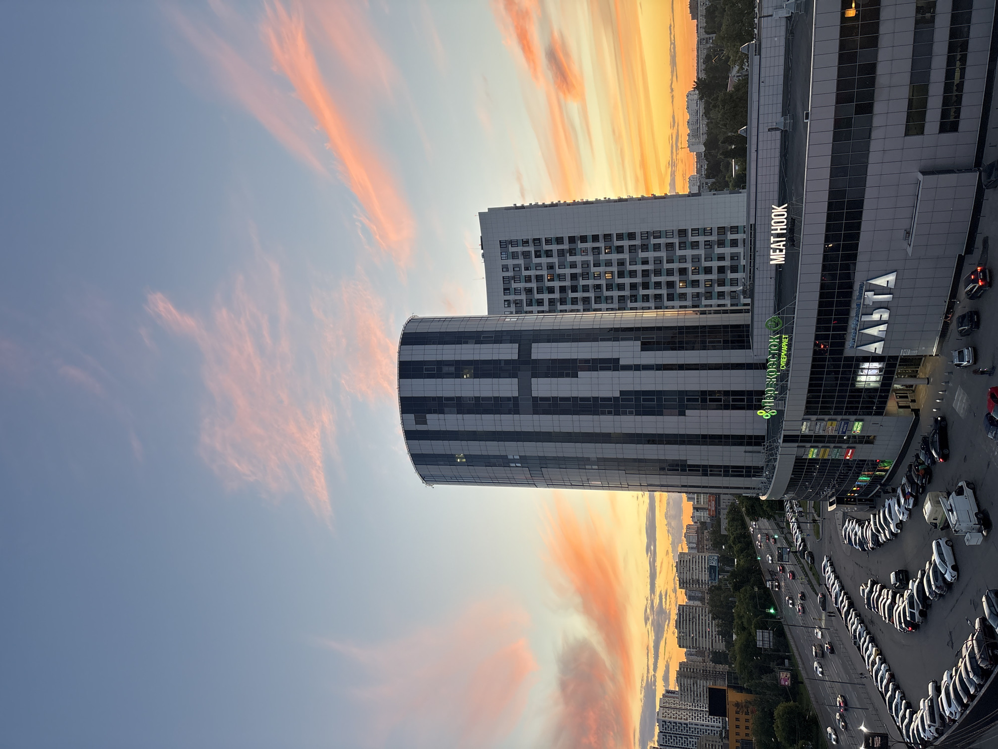 The image captures a city skyline during a sunset. The sky is filled with vibrant orange and pink clouds, suggesting fair weather and the golden hour. In the foreground, a modern, multi-story building with a glass facade dominates the right side of the frame. Above its entrance, illuminated in white neon letters, is the text "MEAT HOOK". Below this, in green, is text that appears to say "супермаркет" (supermarket) and possibly a logo. To the left of this building is another tall, cylindrical building with a dark and light striped facade. Further back, a row of residential buildings can be seen.
The image also shows a street with several cars driving along it, and a large parking lot filled with many cars, mostly white, parked in neat rows. The presence of cars and a supermarket suggests a commercial or residential urban area. The overall mood is serene, enhanced by the beautiful sunset colors. The photograph appears to be taken from an elevated position, looking down on the scene.