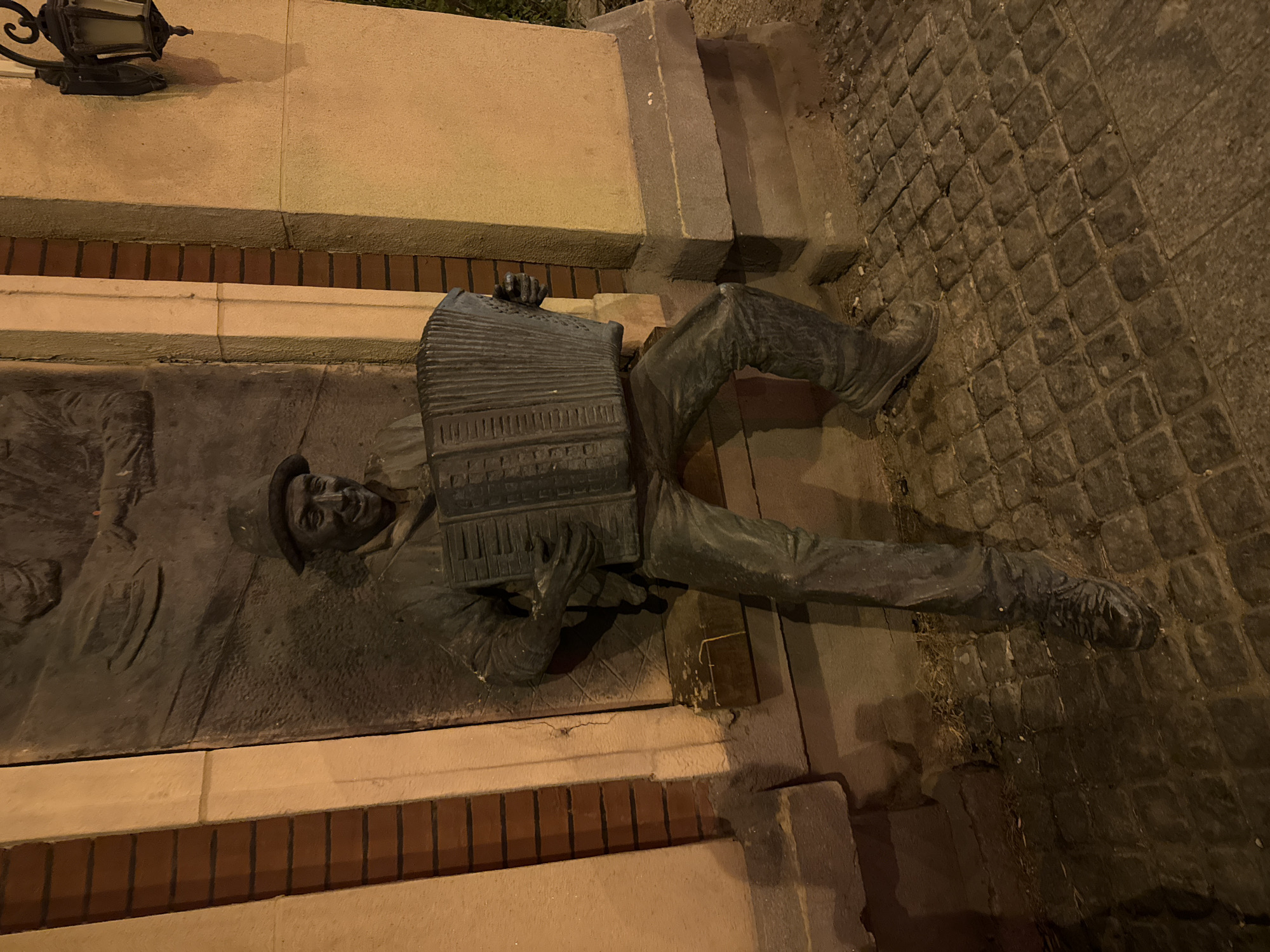 This nighttime image, captured in Shenyang City, China, displays a unique public art installation. The central subject is a bronze-colored sculpture of a man, seemingly emerging from a dark, rectangular panel integrated into a multi-layered wall. The man is depicted playing an accordion, with his upper body and instrument as a high-relief sculpture, while his legs and feet are fully three-dimensional, extending onto the ground. He wears a hat, jacket, and pants, and his expression suggests a cheerful or focused musical performance.
The wall behind the sculpture features a light-colored, smooth upper section, a distinct horizontal band of reddish-brown bricks, and a light-colored base. To the left, a traditional, ornate dark metal lantern is mounted on the wall, providing artificial illumination that casts strong shadows and highlights across the scene. To the right of the figure, several concrete steps ascend. The ground level, where the man's feet are placed, is paved with dark, irregularly shaped cobblestones. The overall lighting suggests it is late evening or night, with the artificial light source creating a dramatic atmosphere. No discernible text is visible within the frame.