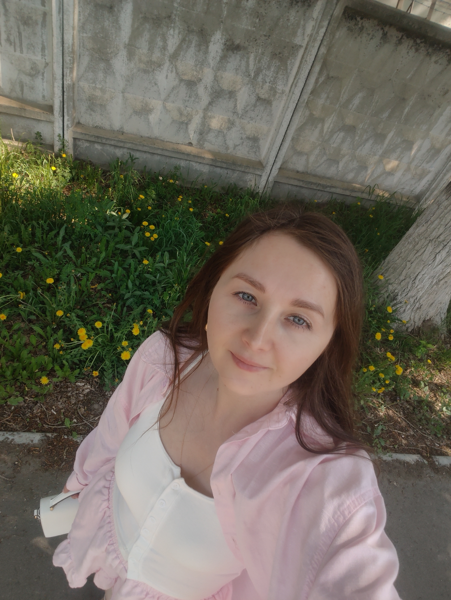 The image shows a woman in an outdoor setting. She is wearing a white top and a light pink button-down shirt. The background includes a concrete wall, grass, and yellow dandelions. A tree trunk is also visible on the right side of the image. She is holding a small white purse or bag. The location appears to be a roadside or park-like area.