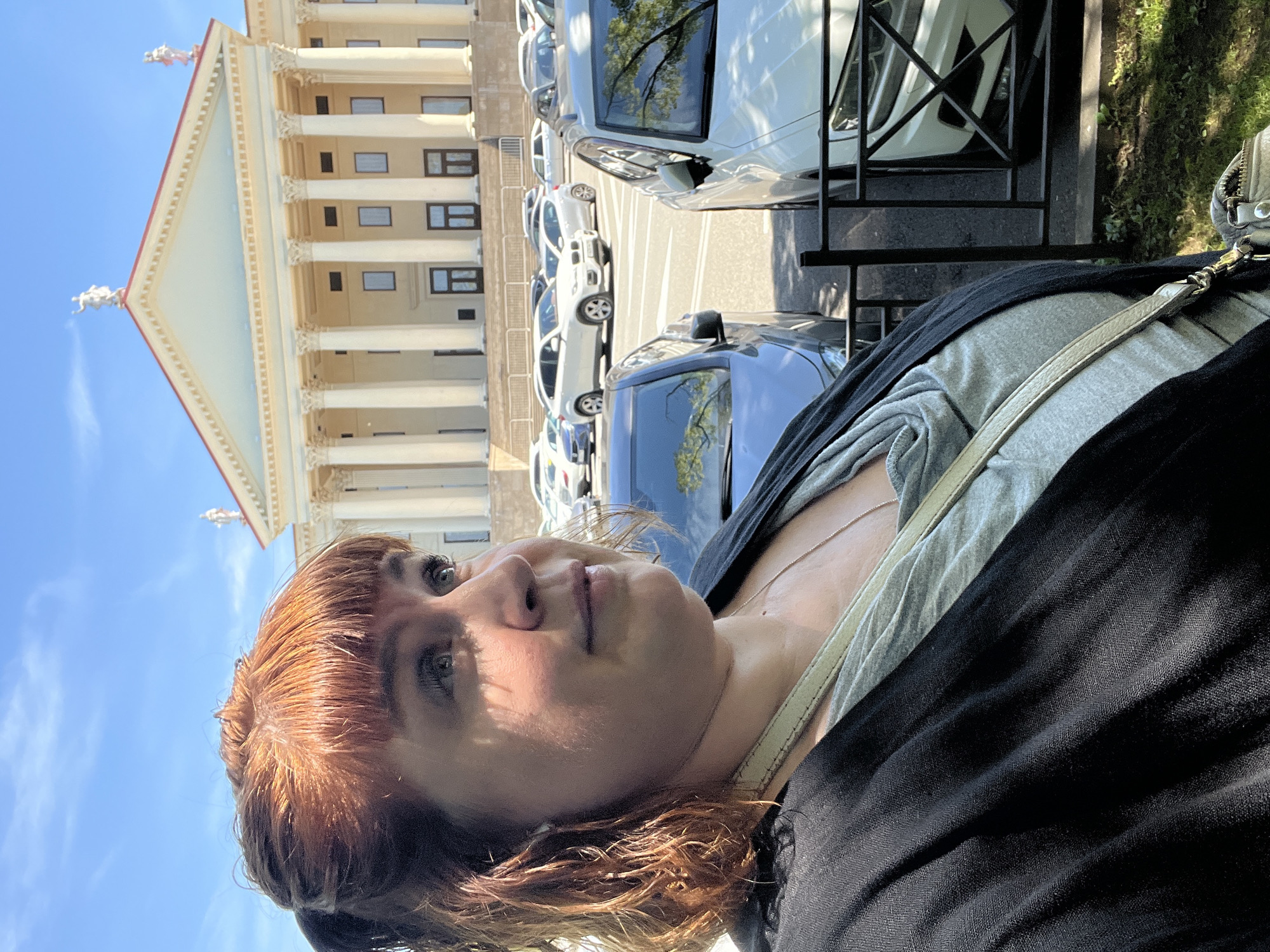 A close-up, upward-facing shot captures a woman with reddish-brown hair and light eyes in the foreground, occupying the left half of the frame. Her face is partially shaded by strong sunlight. She wears a grey top and a black outer garment, with a light-colored strap from a bag or purse visible across her shoulder. Her gaze is directed upwards and to her right, beyond the immediate foreground.
In the mid-ground, a parking area with several cars is visible, including white, silver, and a blue vehicle closer to the viewer. Behind the cars stands a majestic, light-colored classical building, characterized by its imposing triangular pediment and numerous tall columns with ornate capitals. Two small, white statues or figures are perched on the roofline at the base of the pediment. The building features several rectangular windows.
The sky above is a clear, bright blue, indicating a sunny day. Shadows suggest a high sun, likely midday or early afternoon. A black metal railing or fence separates the foreground from the cars. The architectural style of the building points to a grand public structure, consistent with the location context of Sochi, Russia. No visible text is present in the image.