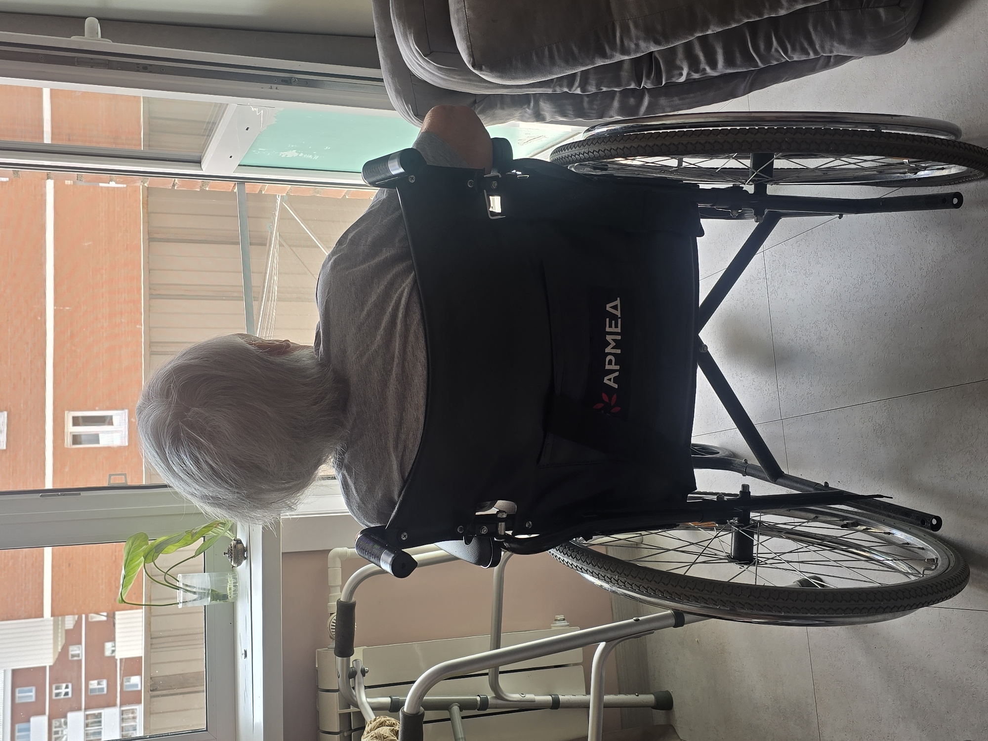 The image depicts a rear view of an elderly person with gray hair sitting in a black wheelchair, facing a window. The person is wearing a gray t-shirt. The wheelchair has the brand "APMED" visible on the backrest. To the left of the wheelchair, a walker is positioned near a radiator. On the windowsill, a plant in a glass of water and a small decorative object are visible. Outside the window, brick buildings and a fence can be seen. The scene appears to be indoors, possibly a room in an apartment or care facility. The location is identified as Ulan-Ude, Russia.