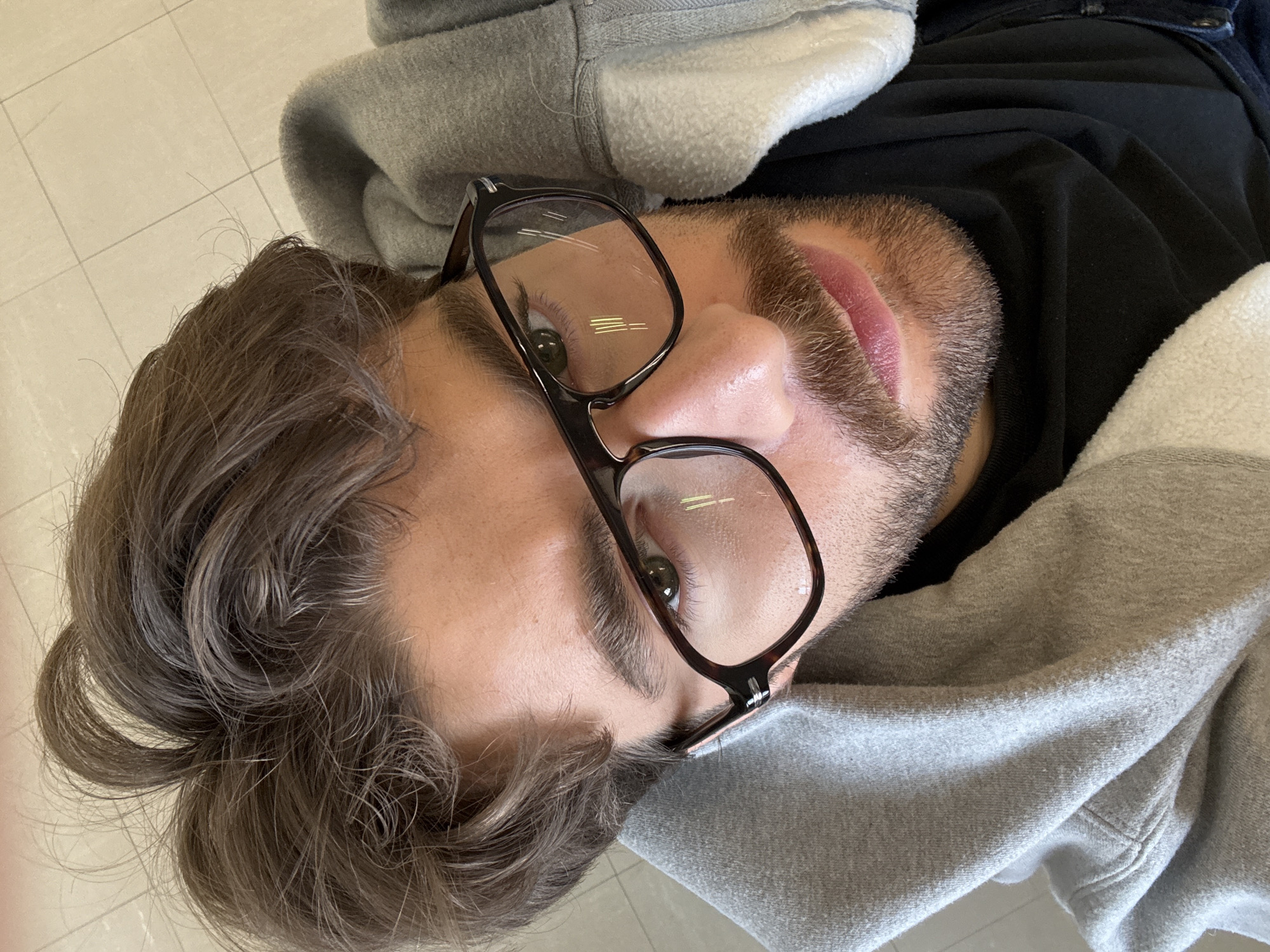 A close-up, top-down portrait captures a person, likely male, lying down and looking directly into the camera. The individual has dark brown, wavy hair with visible lighter or grey strands, particularly around the temples. Thick, dark eyebrows frame greenish-brown eyes, which are seen through a pair of dark brown, rectangular eyeglasses, possibly with a tortoiseshell pattern. Reflections of linear light fixtures are prominent on the lenses.
The person sports a short, dark brown beard and mustache. Their lips are full and reddish-pink. They are dressed in a black hooded sweatshirt with a gray and white fleece-lined hood and collar area. The gray fabric appears soft and textured. A fragment of blue material, possibly denim, is visible near the person's shoulder.
The background consists of light-colored, off-white or beige tiled flooring with visible grout lines, suggesting an indoor setting. The person appears relaxed, posing for the photograph. No specific activity is occurring beyond being photographed while resting. There is no visible text in the image, and no cues to determine the time of day or weather are present.