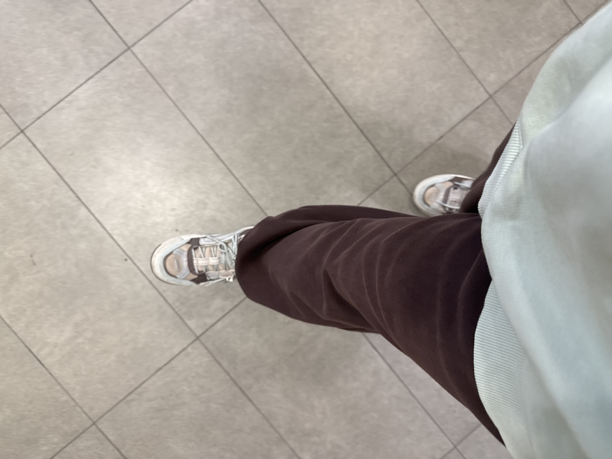 The image shows a close-up, top-down view of a person's legs and feet. The person is wearing brown sweatpants and light blue and white sneakers with laces. The feet are positioned on a tiled floor with a grid pattern. The environment appears to be indoors, possibly a public space like a shopping center or a school hallway, given the clean, uniform tiling. The lighting is even and neutral. There is no indication of the specific location within Lyubertsy, Russia, as the image lacks any recognizable landmarks or context clues. The scene is static and shows no activity other than the implied presence of the person.