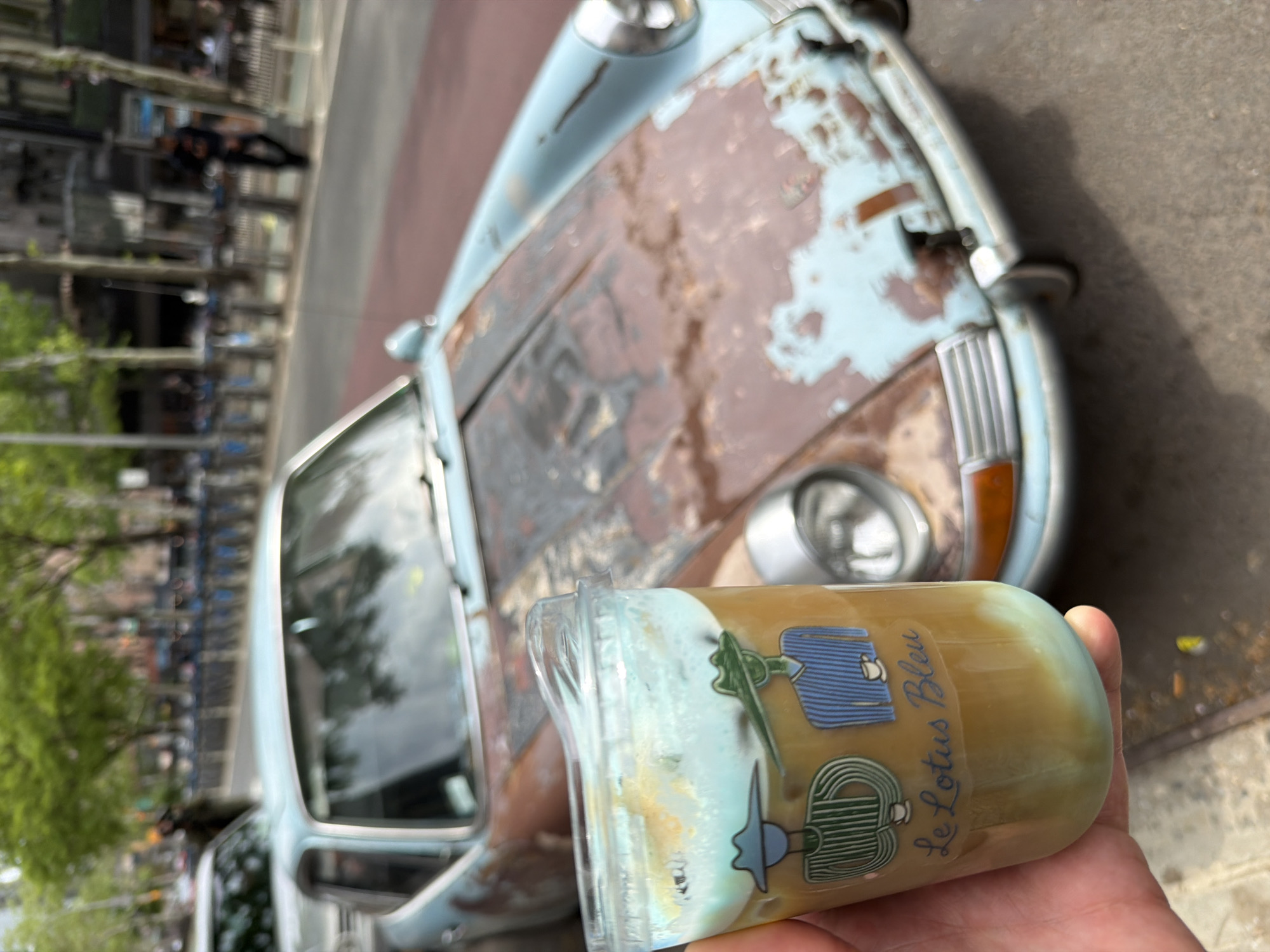 A person holds a glass of Le Lotus Bleu cafe in front of a light blue, rusted Volkswagen Beetle in New York City, United States. The scene features a blend of urban and automotive elements. The Beetle, with its worn and rusted exterior, adds a vintage touch to the setting. The glass, labeled with "Le Lotus Bleu," appears to contain a layered beverage. In the blurred background, the urban landscape includes trees and buildings.
