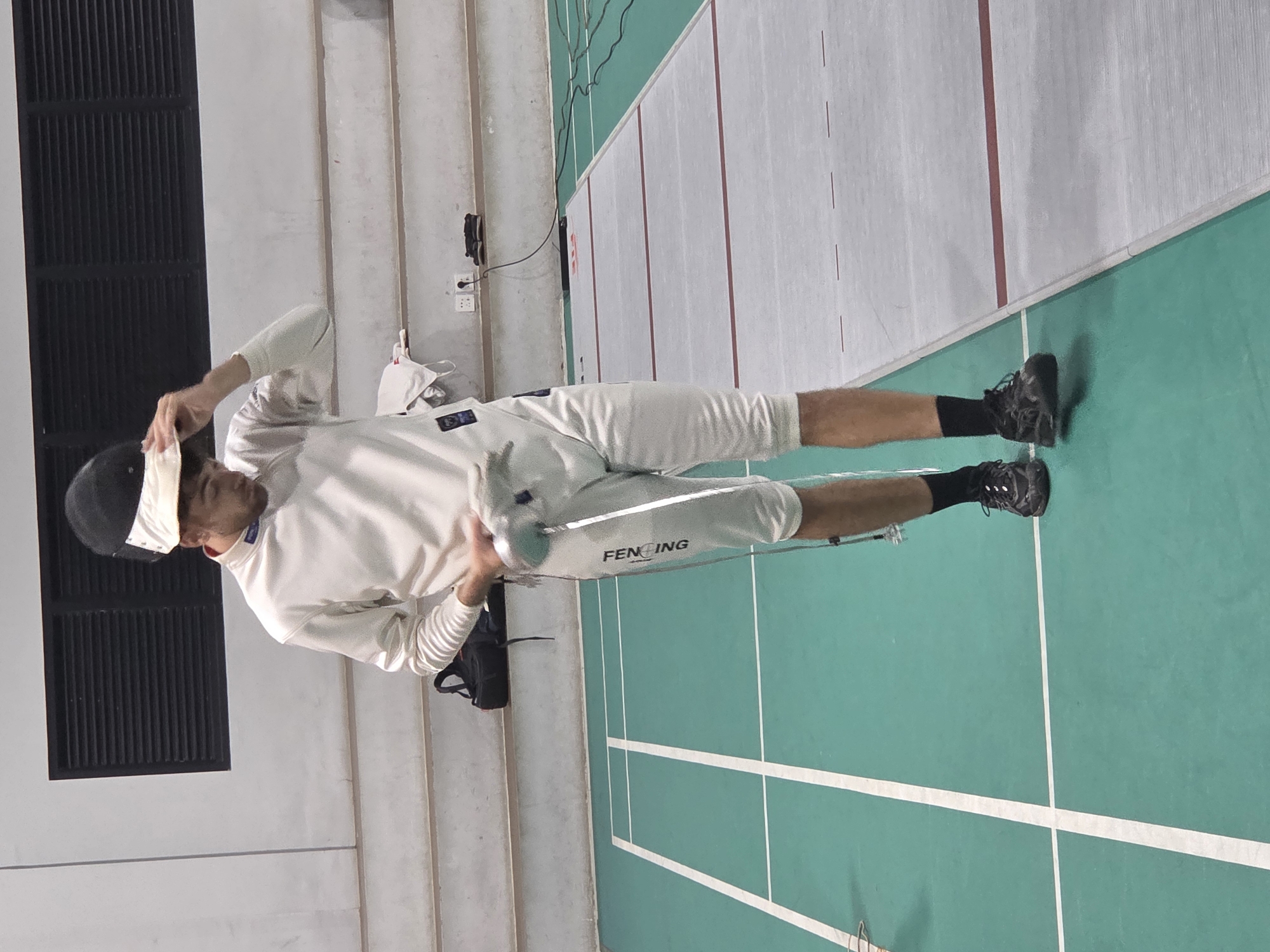 A male fencer is shown in a sideways view, adjusting his fencing mask in an indoor fencing hall. The hall has a green fencing strip with white boundary lines, and a gray area next to it. There are spectator stands in the background, along with electrical outlets and a hanging equipment bag. "FENCING" is visible on the fencer's pants. The fencer is wearing a white fencing uniform and black fencing shoes.
The image was likely taken in North Kuta, Indonesia.