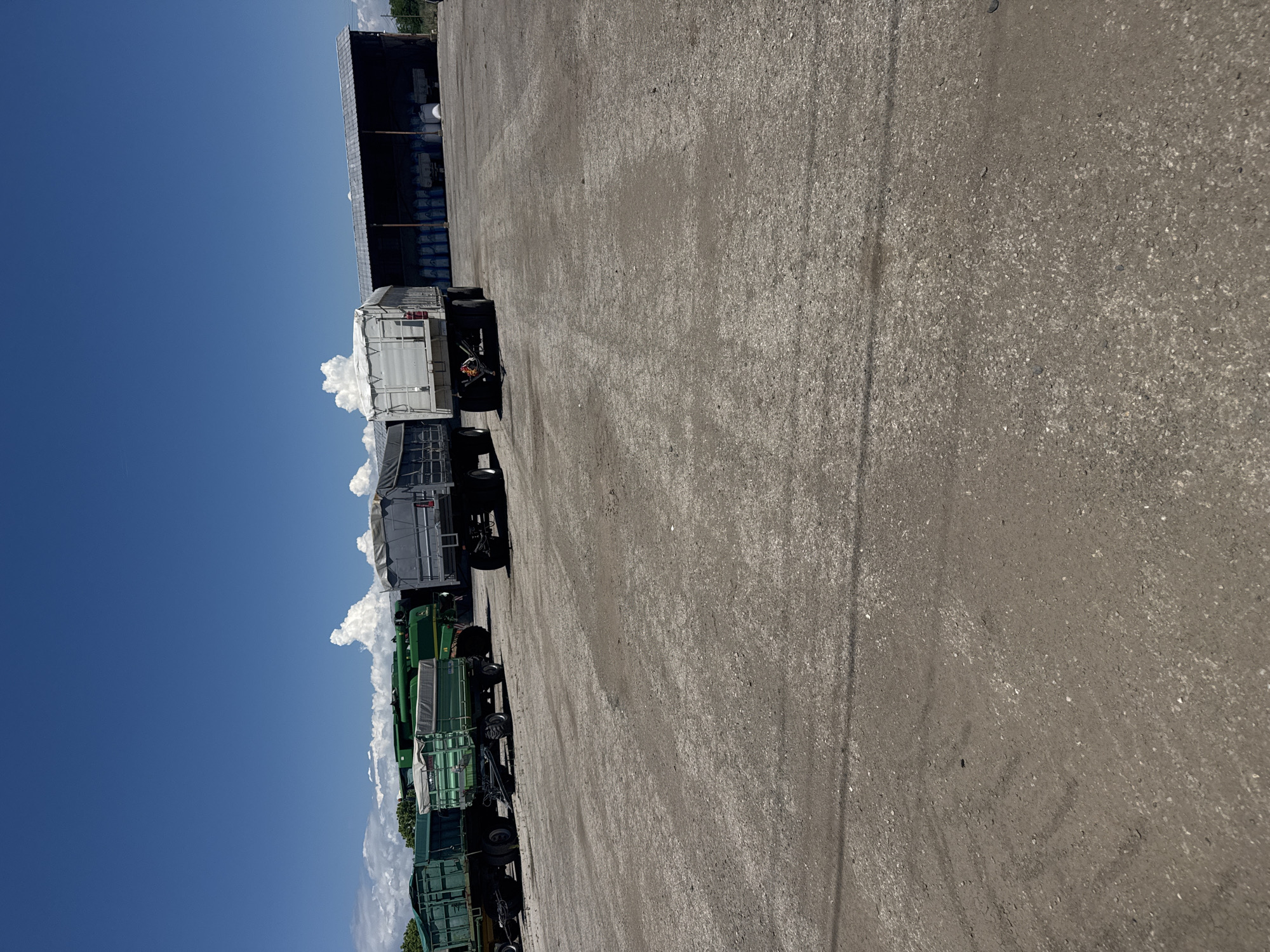 The image shows a row of vehicles parked on an asphalt lot under a bright blue sky. The vehicles include two white trailers and several green agricultural trailers. A large industrial building with visible stored goods is partially visible in the background. The location is potentially Sarnevo, Bulgaria, judging by the metadata. The scene appears to be a storage or parking area for agricultural or transport equipment.
