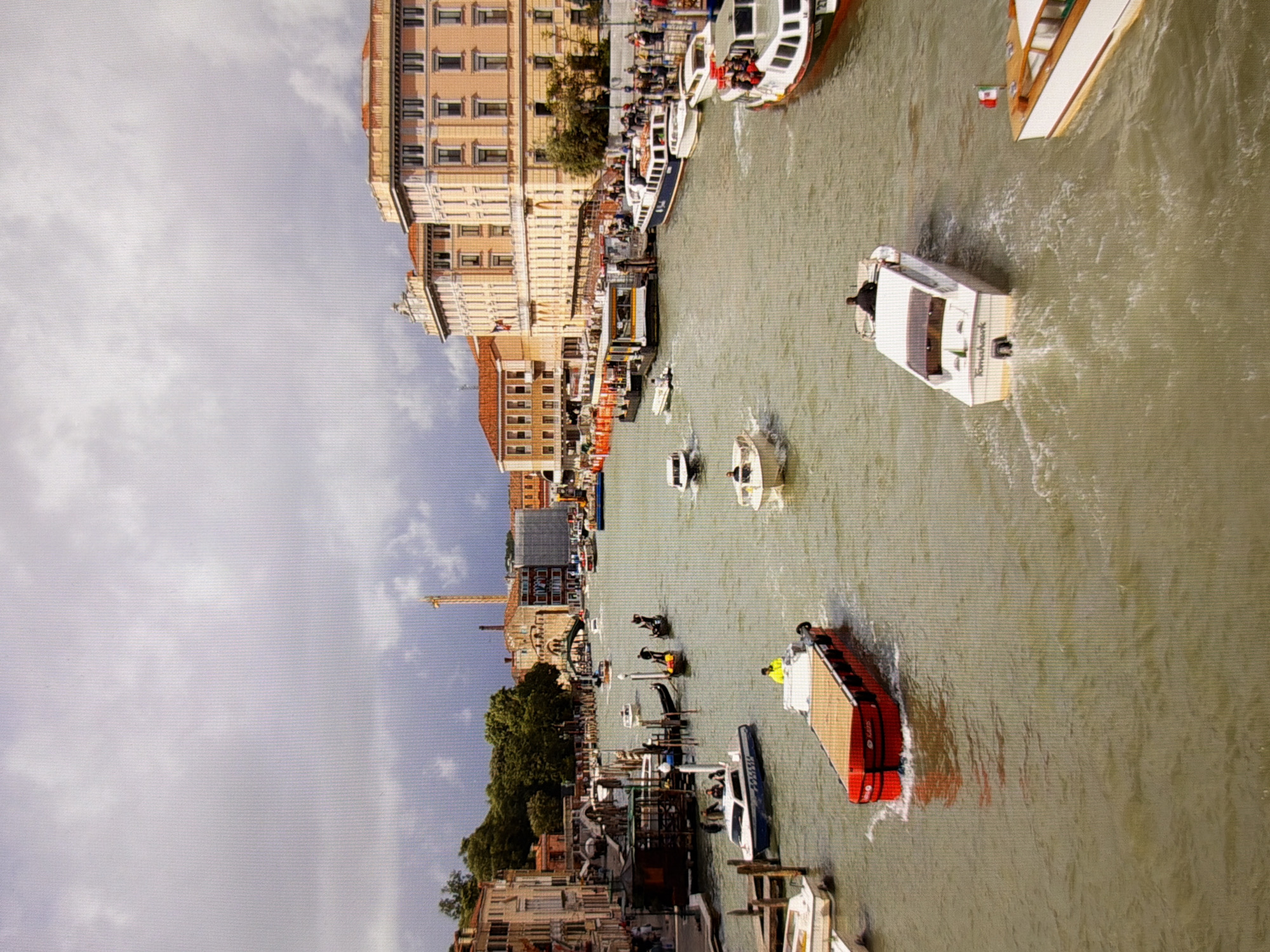 This is a wide-angle, eye-level shot of a canal in Venice, Italy, on a cloudy day. The canal is bustling with various boats, including a red barge with a wooden deck, a white police boat, several smaller white speedboats, gondolas, and a larger white and blue passenger ferry. People are visible in some of the boats, operating them or as passengers, and on the banks of the canal, some walking and some standing on a pier. Buildings line the canal, featuring a mix of architectural styles, some with terracotta roofs and others with beige facades. A construction crane is visible in the distance. The water is a murky greenish-brown, with visible wakes from the moving boats. The overall impression is a busy urban waterway with historical architecture and various modes of transport.