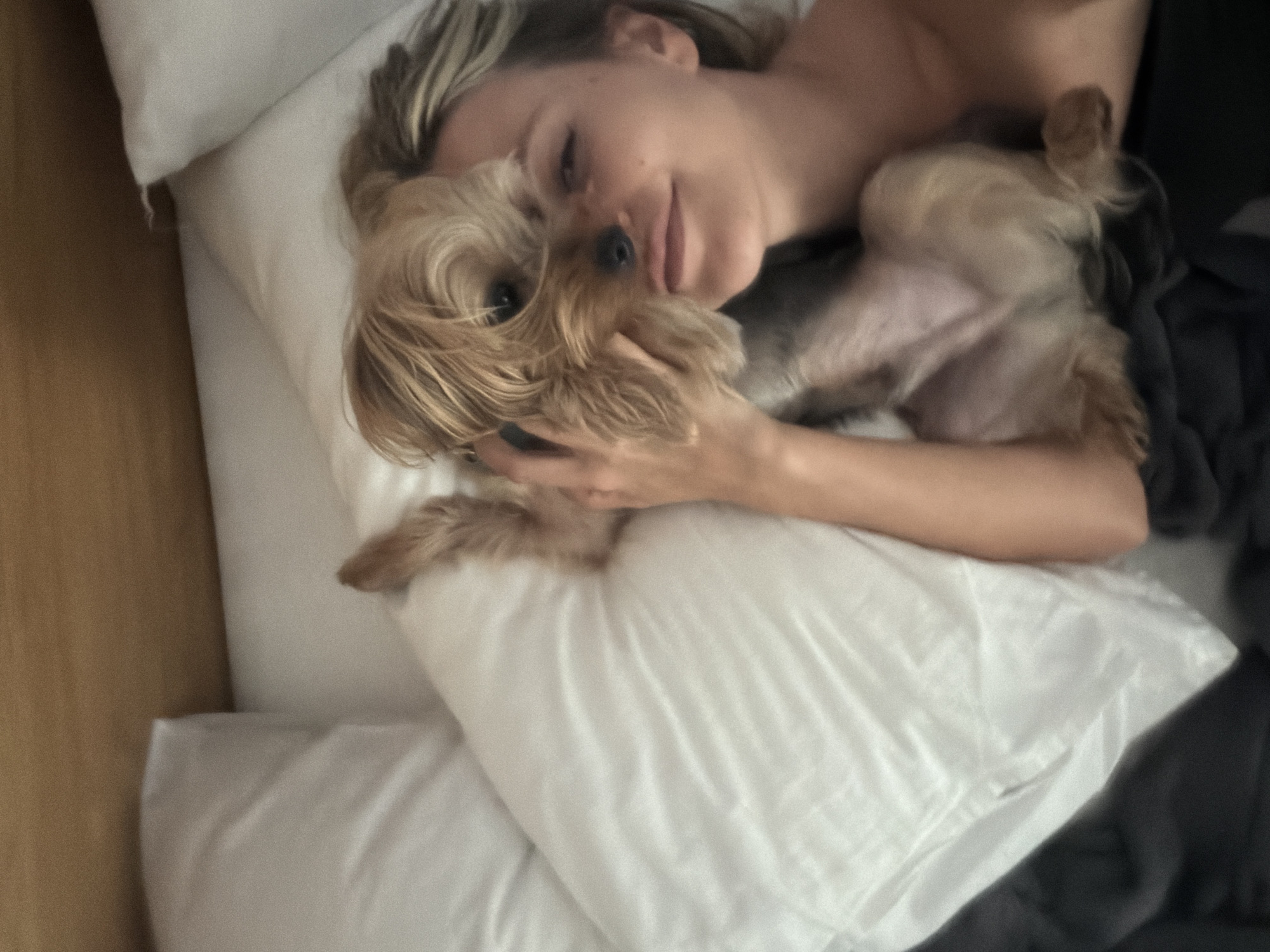 A person and a small dog are lying in a bed, with their heads close together. The person's eyes are partially closed, and they appear to be smiling. The dog, a Yorkshire Terrier, has its head resting on the person's cheek. The bed is made with white linens. A wooden headboard is visible in the background. The scene is lit by natural light. The location in the city of Thep Krasatti, Thailand is not discernible from the image.