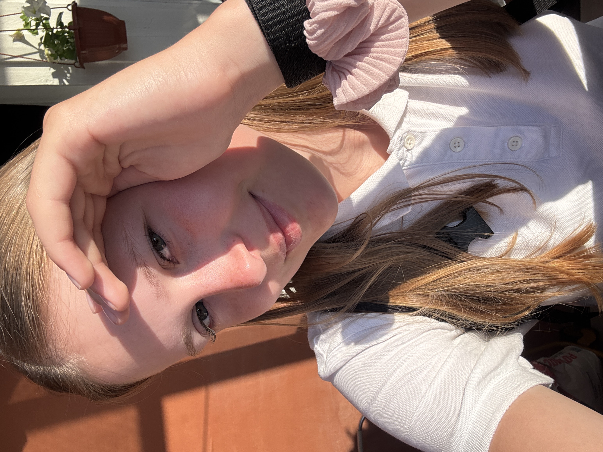 A young person with light brown hair and fair skin is holding their hand to their forehead, looking at the camera. They are wearing a white polo shirt and a black wristband with a pink scrunchie. The background is a blurry outdoor scene with a brown surface and hints of greenery, including a potted plant on a white railing. The photo was not taken in Chaikovsky, Russia.