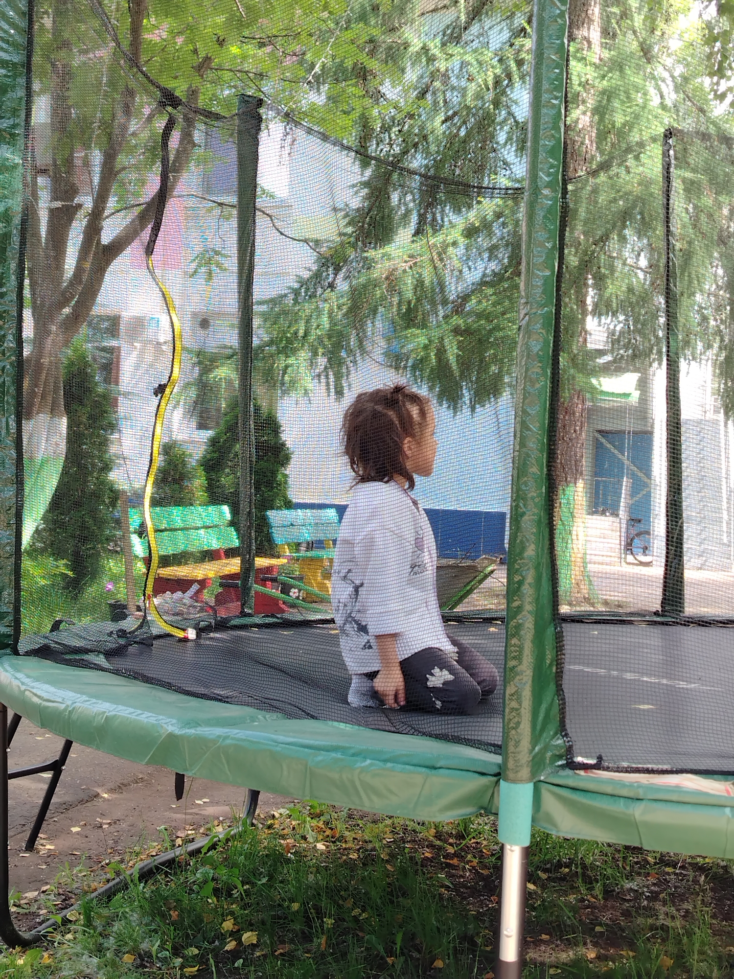 A young child, seen from the side and from behind, sits on a trampoline inside a safety net. The child has dark hair pulled back in a ponytail and wears a white shirt with a black design and dark pants. The trampoline has a green padded edge and a black jumping surface. In the background, through the net, there are trees, a building with blue and white sections, green benches, and a bicycle. The scene appears to be outdoors, possibly in a residential area or park, with dappled sunlight. The location is identified as Chaikovsky, Russia.
