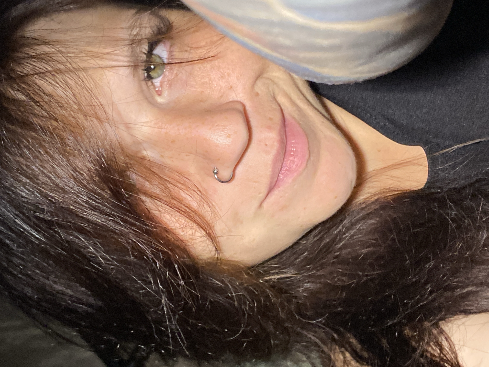 The image is a close-up, low-angle shot of a young woman with long, dark brown hair that partially covers her face. She has fair skin with freckles and is wearing a silver hoop nose ring. Her eyes are light green, and she is smiling faintly. She is wearing a black top. A silver, cylindrical object is visible on the right side of the frame, partially obscuring her face. The background is dark and indistinct. There is no discernible indication of the city of Chaikovsky, Russia, in this image.