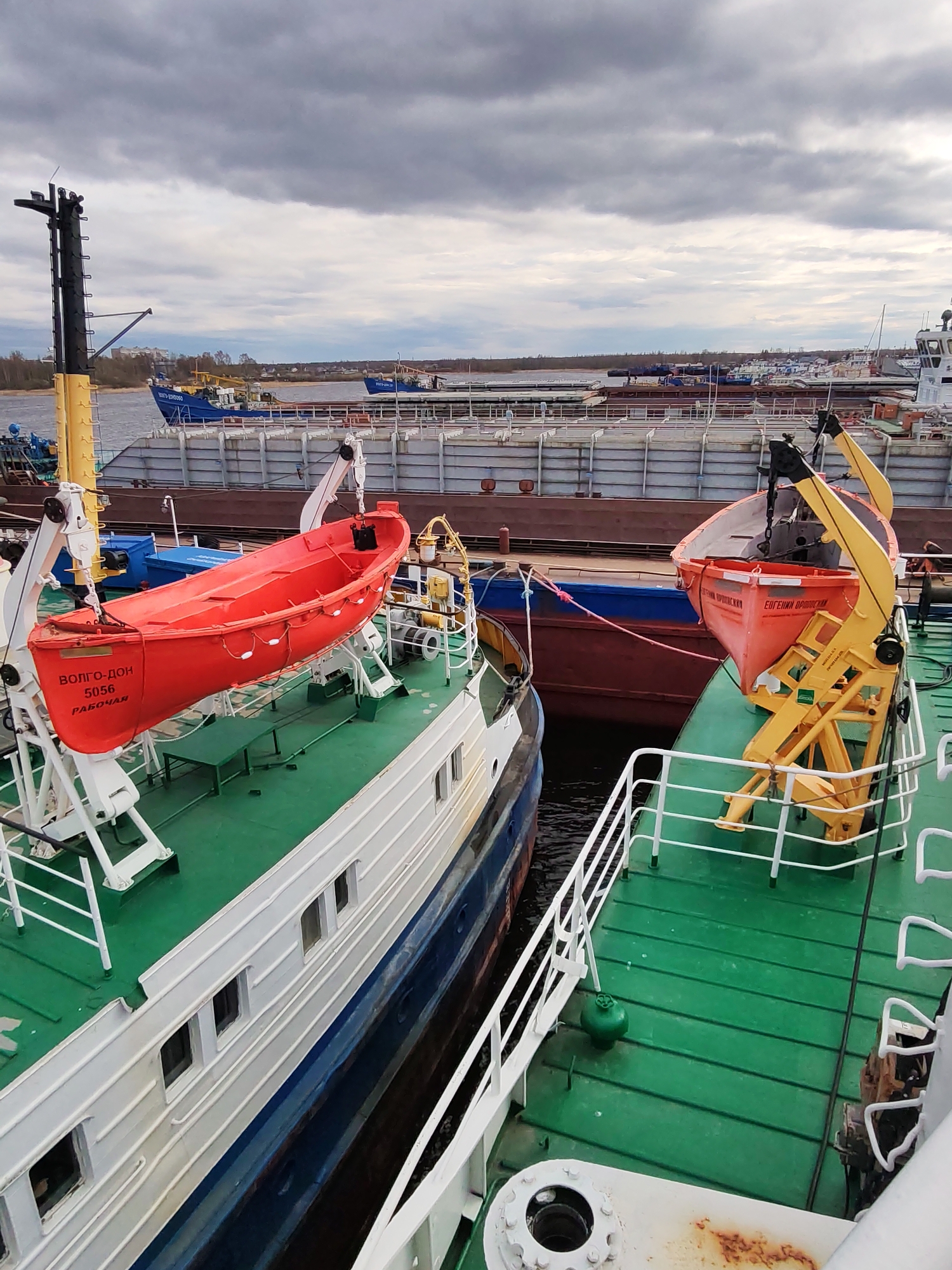 The image depicts a view from the deck of one ship towards other docked vessels in what appears to be a port or shipyard setting. The scene is dominated by the green decks of the ships and two orange lifeboats. One lifeboat has the Russian text "ВОЛГО-ДОН 5056 РАБОЧАЯ" on its side. The other says "ЕВГЕНИЙ ОРОДНИКОВ". Various other ships, barges, and structures are visible in the background, as well as the river. The sky is overcast. Based on the visible Russian text on the lifeboats and the reference to Rybinsk, Russia, it is reasonable to assume this scene is located there.