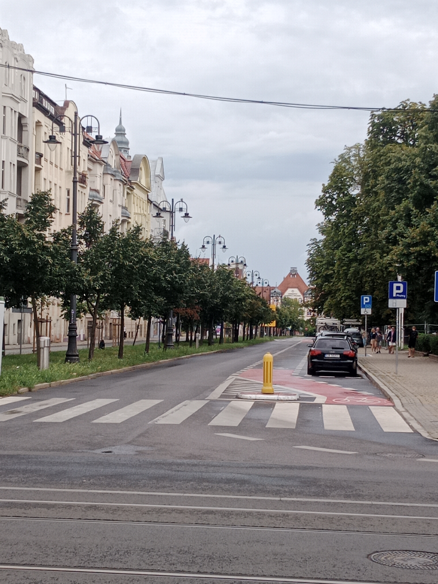 This is a photograph of a street in Bydgoszcz, Poland, taken on a cloudy day. The street is lined with buildings on the left and trees on the right. A crosswalk is visible in the foreground, with a yellow bollard marking its edge. A black car is parked on the right side of the street, near a "P" parking sign. Several people are walking on the sidewalk to the right of the car. The sky is overcast. The architecture of the buildings suggests they are historic. The presence of tram tracks in the lower part of the image indicates that this is a street with public transportation.