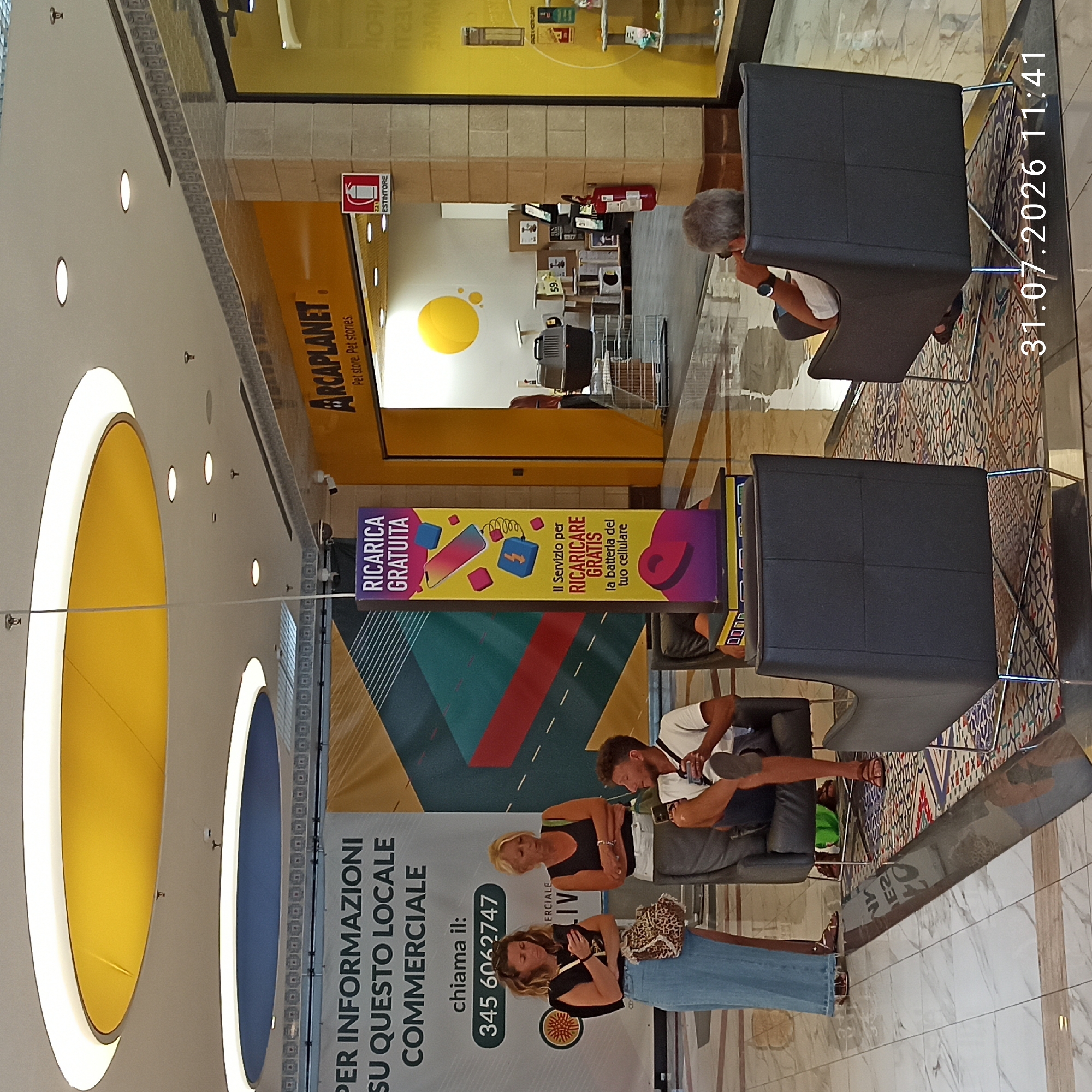 The image captures an interior scene, likely within a commercial establishment. The ceiling features circular lighting elements, one yellow and one blue, with surrounding bright white lights. In the background, a yellow sign with the text "ARCAPLANET Pet store. Pet stories." is visible, alongside an "ESTINTOR" fire extinguisher.
In the foreground, three individuals are present. Two women, one with blonde hair and the other with brown hair, stand looking at their phones. The woman with blonde hair wears a black tank top and white pants, while the woman with brown hair wears a black top and a long blue skirt, carrying a leopard-print bag. A man with curly brown hair sits in a dark grey armchair, also looking at his phone. He is wearing a white t-shirt and dark shorts. He is sitting on an oriental-style rug.
A tall, vertical banner displaying a mobile phone being charged, with the text "RICARICA GRATUITA" (Free Recharge) and "Il Servizio per RICARICARE GRATIS la batteria del tuo cellulare" (The service to recharge your phone battery for free) is positioned between the banner and the store entrance.
To the right, a view into the Arcaplanet store reveals pet supplies, including kennels, shelves with products, and cat trees.
A timestamp "31.07.2026 11:41" is superimposed on the bottom right of the image. The flooring appears to be a combination of light-colored marble tiles and a patterned rug. The overall atmosphere suggests a modern shopping center or mall. The location is identified as Modugno, Italy.