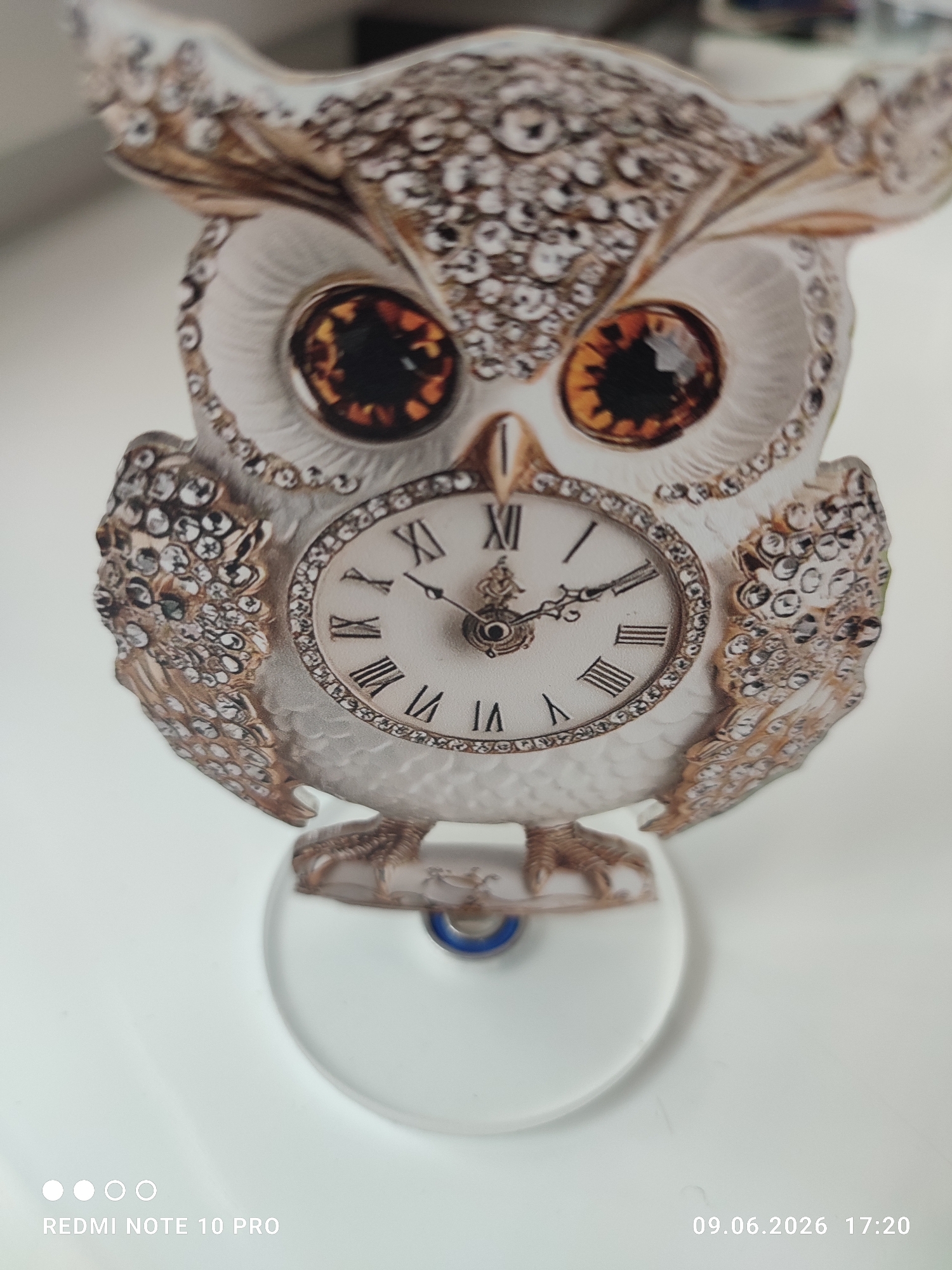 The image shows a decorative owl clock with Roman numerals on its face. The owl is white with gold accents and is adorned with small, sparkling rhinestones. The clock hands are dark and appear to be set at approximately 1:09. The owl is mounted on a clear, circular base. The background is a plain white surface, suggesting an indoor setting. The bottom left corner of the image displays the text "REDMI NOTE 10 PRO" and the bottom right corner shows the date and time "09.06.2026 17:20". The location is not specified, so it is Unknown, Unknown.