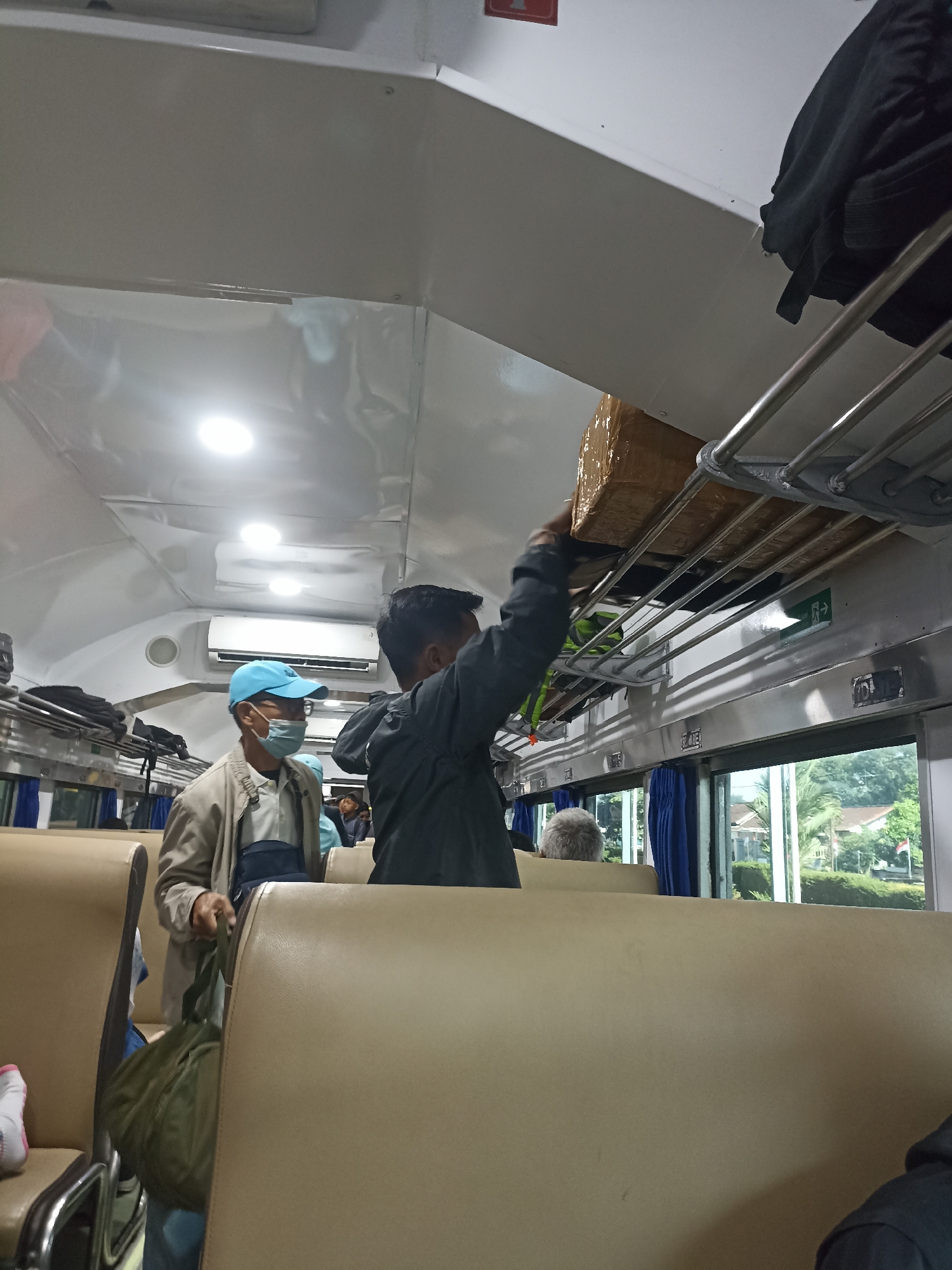 The image depicts the interior of a passenger train carriage, likely in Jambewangi, Indonesia, with passengers and luggage. In the foreground, two men are visible. One man, wearing a light-colored jacket and a blue cap, is also wearing a light blue face mask and carrying a dark green duffel bag. The second man, dressed in a dark jacket, is reaching up to place a large, brown cardboard box onto the overhead luggage rack. The train carriage has beige upholstered seats, chrome luggage racks, and bright interior lighting. Through the windows, a glimpse of greenery and buildings can be seen, suggesting the train is either stationary or moving through a semi-urban or rural area. The overall atmosphere is casual, with passengers engaged in stowing their belongings.