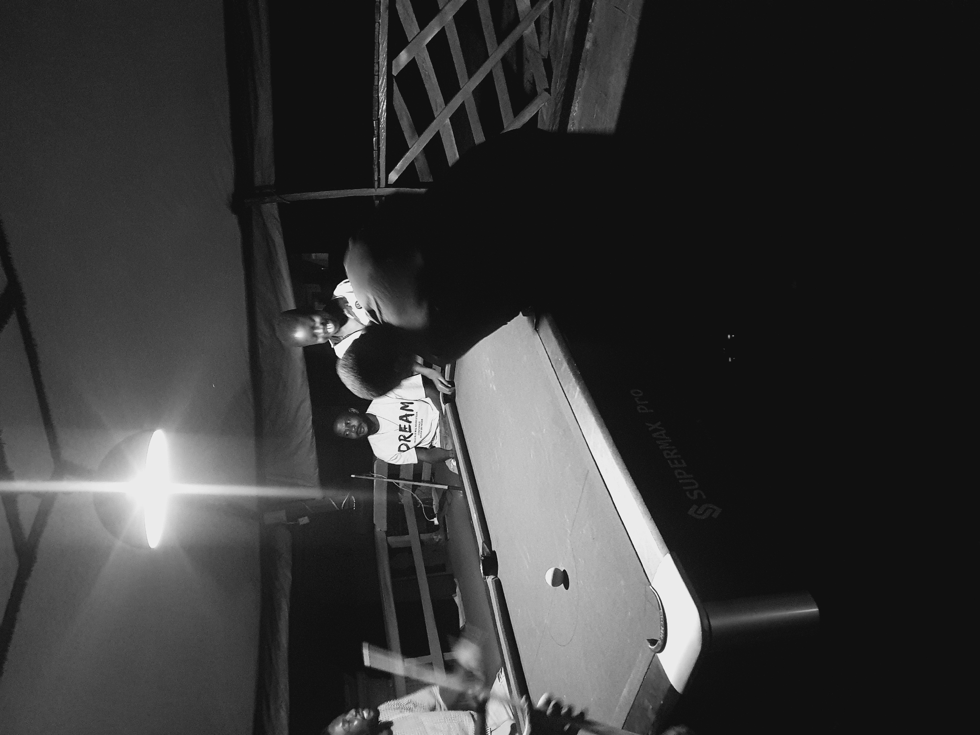 A group of young men are playing pool in an outdoor, dimly lit area, possibly a bar or recreational space, in Lokoja, Nigeria. The scene is captured in black and white, with harsh overhead lighting casting strong shadows. In the foreground, a pool table with a single white ball is visible. The table has "SUPERMAX Pro" written on its side. Two men are actively engaged with the game. One man, wearing a t-shirt with "DREAM" printed on it, is seated and looking towards the table. Another man, whose face is mostly obscured by shadow, is bent over the table, appearing to be preparing a shot. In the background, other individuals are present, some looking towards the game. The environment appears to be partially covered by a roof or awning, with wooden railings and supports visible. The overall atmosphere suggests a casual, social gathering.