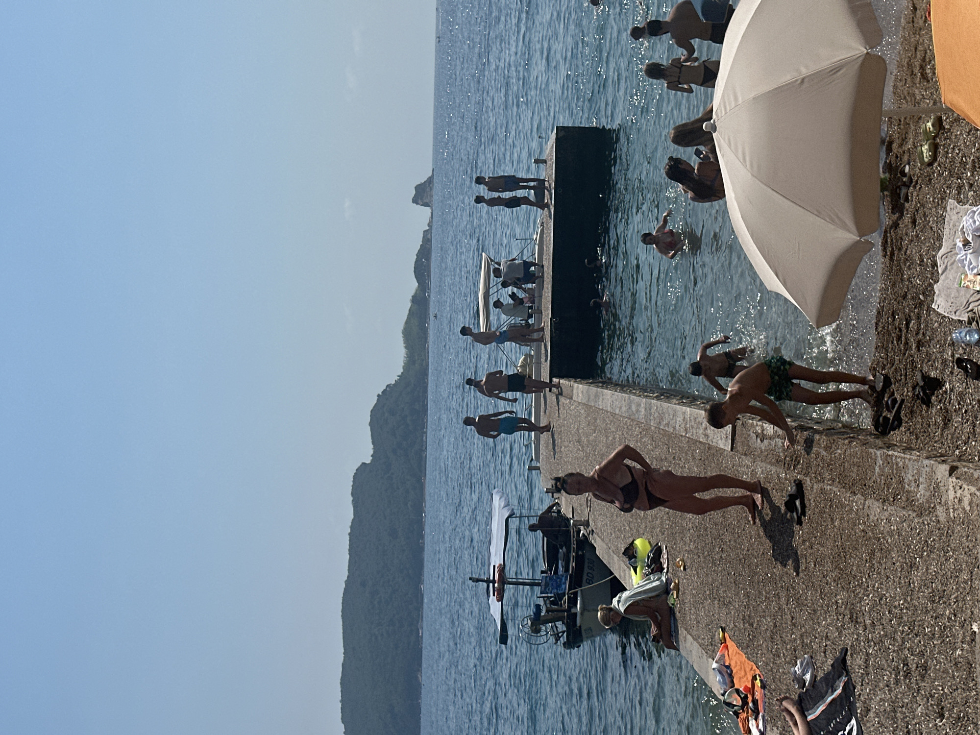 The image depicts a sunny day at a pier in Budva, Montenegro. Several people are enjoying the water and the pier. In the foreground, a woman in a black bikini stands on the pier, facing the camera. To her left, a young boy in green shorts is entering the water. A large, light-colored umbrella is positioned in the lower right corner, shading a section of the pebble beach.
The pier extends into the clear blue sea, with a small boat docked on its left side. On the pier, various individuals are visible: a woman in a swimsuit is seated with a yellow float, and further down the pier, a group of people are gathered, some standing and some seated. More people are swimming and wading in the water. In the background, a lush, green hillside with a rocky peak rises from the sea, characteristic of the Budva coastline. The sky is a clear, bright blue.