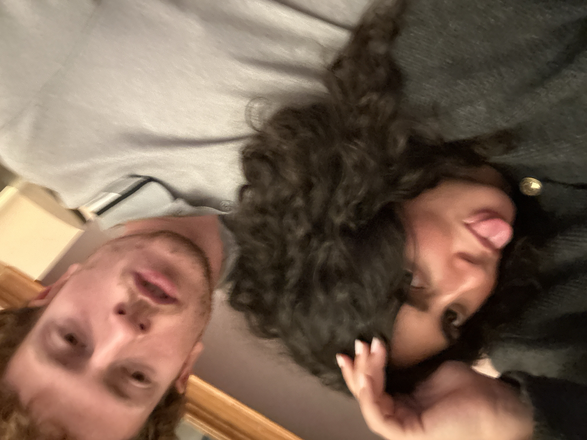 A close-up, rotated image shows two people lying down. On the left is a man with light skin and short brown hair. He is wearing a light gray t-shirt. On the right is a woman with dark curly hair and dark skin, sticking her tongue out. She is wearing a dark-colored shirt with a gold button. The environment appears to be indoors, with a light-colored wall and a wooden trim visible. It is impossible to discern a specific location in New York City from this image.