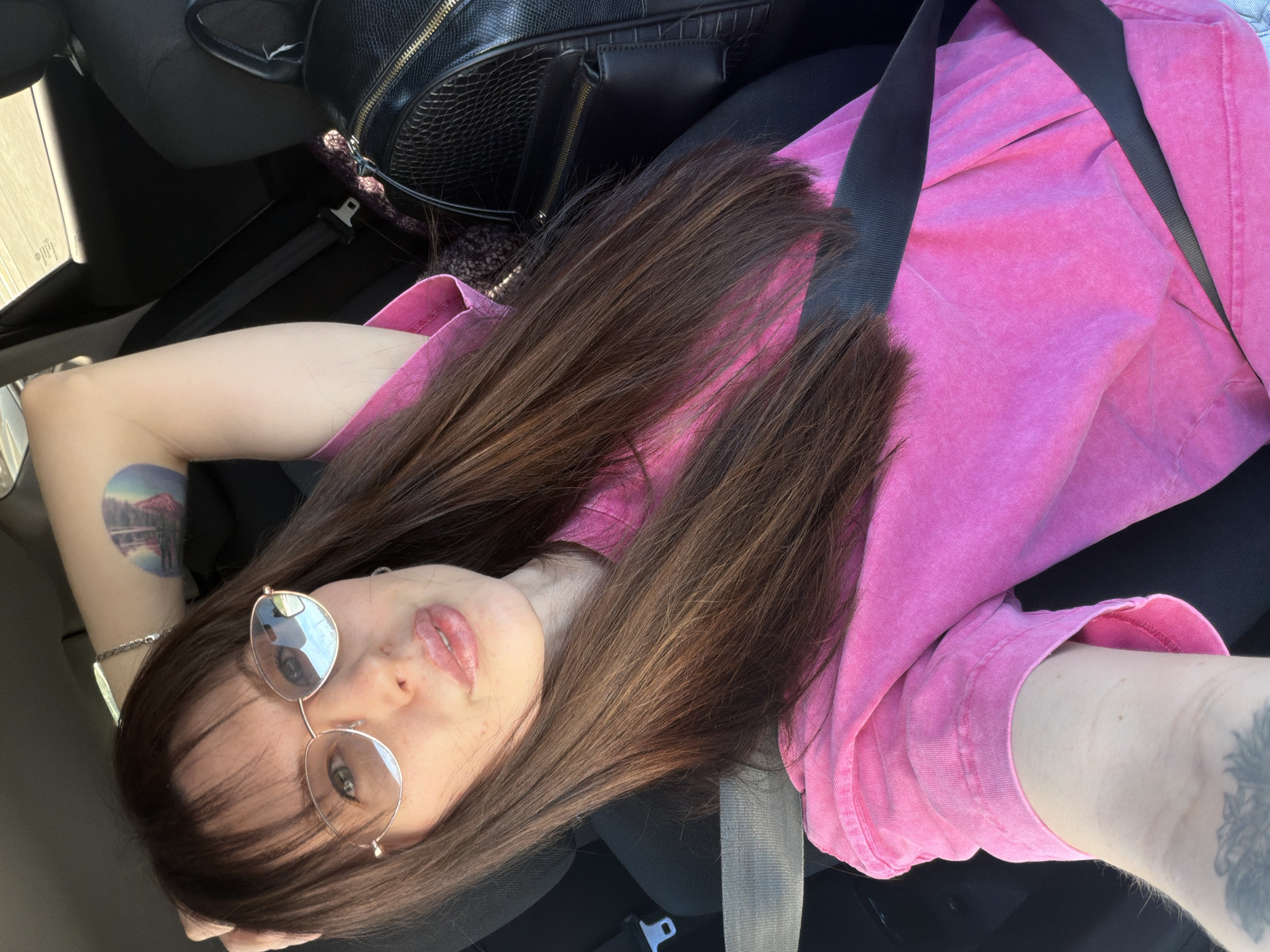 A young woman is inside a car wearing a pink shirt, black seatbelt, and glasses. A black purse sits next to her. She has tattoos on her arms and long brown hair. The location is unknown and cannot be inferred from the given information.