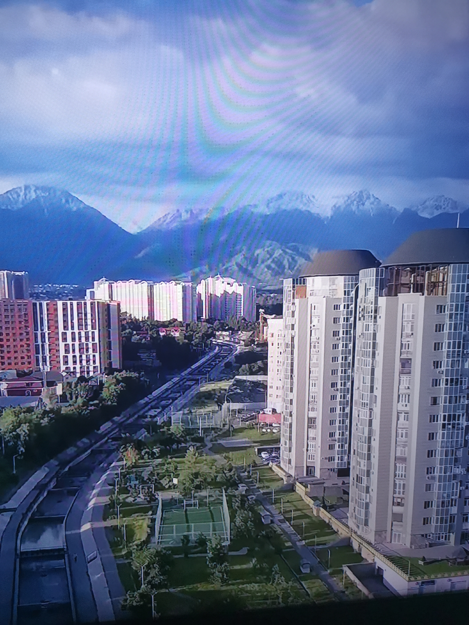 This is an aerial view of a modern city with mountains in the background. The scene features several tall residential buildings, some with rounded, domed roofs, and others with straight, modern facades. There are also lower-rise buildings, lush green trees, and what appears to be a road or elevated railway cutting through the urban landscape. A sports field or court is visible in the green space between the buildings. The sky is partly cloudy, with large, wispy clouds casting shadows over the landscape, suggesting it might be a bright but not entirely sunny day. The lighting and cloud patterns could indicate midday or early afternoon. The presence of snow-capped mountains in the distance provides a striking contrast to the urban development, suggesting a location where mountains are in proximity to a city. There is no visible text in the image. The overall impression is one of a developed urban environment situated in a scenic, mountainous region.