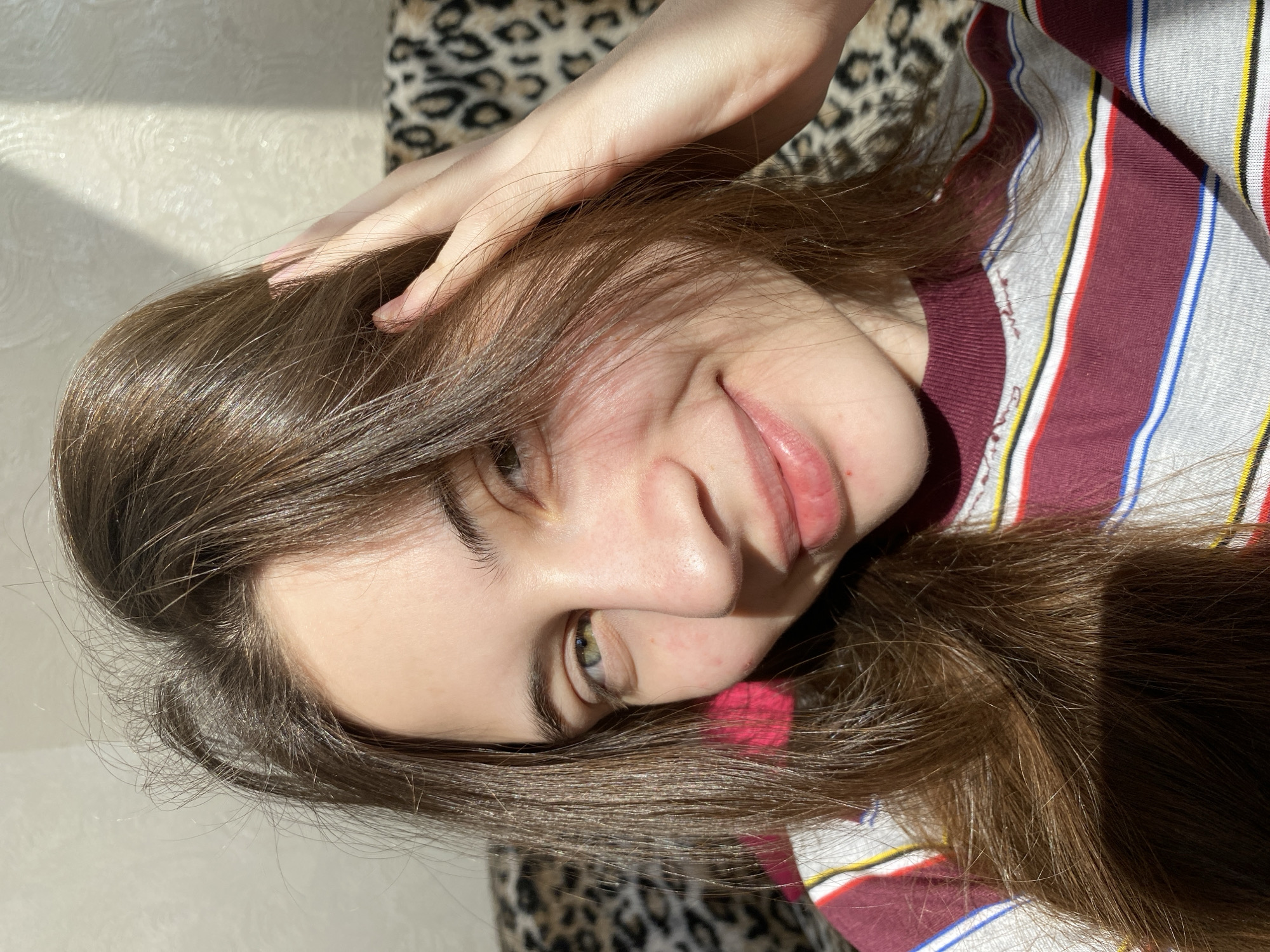This media file presents a close-up, overhead portrait of a young person, likely female, captured indoors in Votkinsk, Russia. The subject is lying down, with their head tilted, looking towards the camera with a subtle, playful smile. Long, glossy brown hair flows around their face, with one hand featuring neatly manicured nails resting on their head, pushing some hair back.
The person has fair skin, with visible green or hazel eyes and a few small reddish spots on their cheeks and nose. Their lips are naturally pinkish. They are wearing a striped t-shirt with horizontal bands in burgundy, grey, white, blue, yellow, and red. Faint, stylized cursive text is subtly visible on one of the burgundy stripes, though it is indecipherable.
The background consists of a light-colored, textured wall and a soft furnishing with a leopard print pattern, suggesting a comfortable, indoor setting such as a bedroom or living room. Bright natural sunlight streams in from an unseen window, casting distinct shadows and highlighting the person's hair and features, indicating it is daytime. The overall impression is one of casual relaxation.