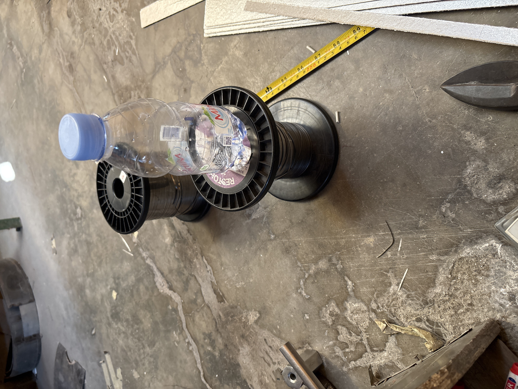 A clear plastic water bottle with a blue cap sits atop a spool of dark wire. The bottle is partially filled with water. The spool of wire is resting on a larger spool of dark wire, both appearing to be made of plastic. To the right of the spools, a yellow measuring tape is laid out on the concrete floor. The tape shows markings in inches and centimeters. Several thin, metallic-colored objects are scattered on the floor, possibly cut pieces of wire. In the background, the concrete floor is uneven and shows signs of wear and discoloration. To the far left, a metallic object with a bolt and nut is visible. The overall scene suggests a workshop or industrial environment. The image does not provide any specific details to determine the location within Telagamurni, Indonesia.