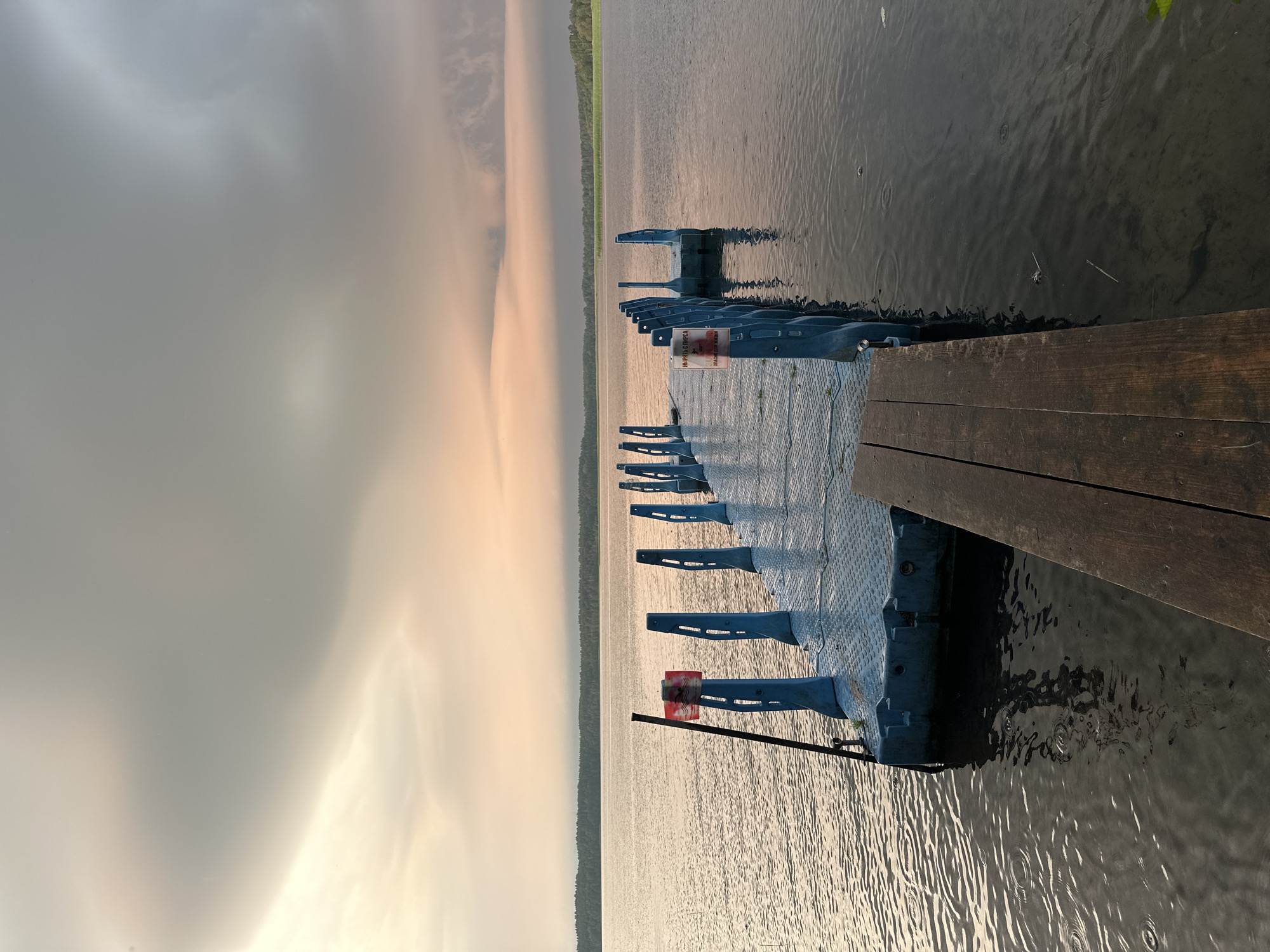 This image depicts a floating pontoon dock extending into a body of water, likely a lake or river, with a treeline visible in the distance. The sky is overcast with dramatic clouds, tinged with the colors of a sunset or sunrise. The water is calm with ripples, suggesting either light rain or wind. The pontoon, made of blue modular plastic sections, has a metal surface and is equipped with blue vertical posts along its length. A small red and white sign is attached to one of the posts, bearing text in Cyrillic that likely warns against diving. In the foreground, a section of a wooden dock is visible. The overall scene is set in a natural environment.