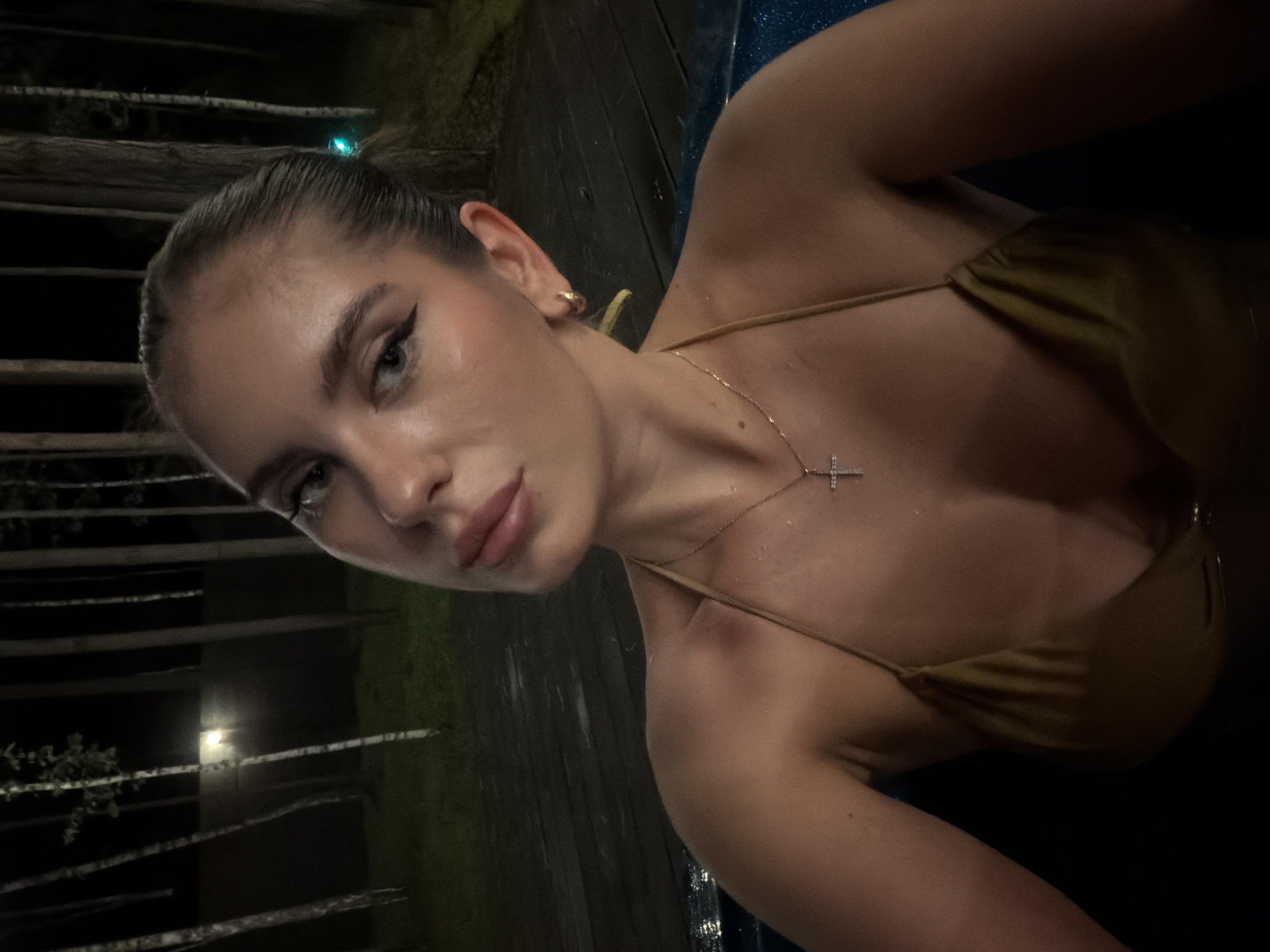 A young woman with dark hair pulled back, wearing a gold bikini top and a cross necklace, is pictured in a hot tub at night. The background shows trees and a wooden deck. A light is visible in the distance behind the trees. The image appears to have been taken in a wooded area near a body of water.