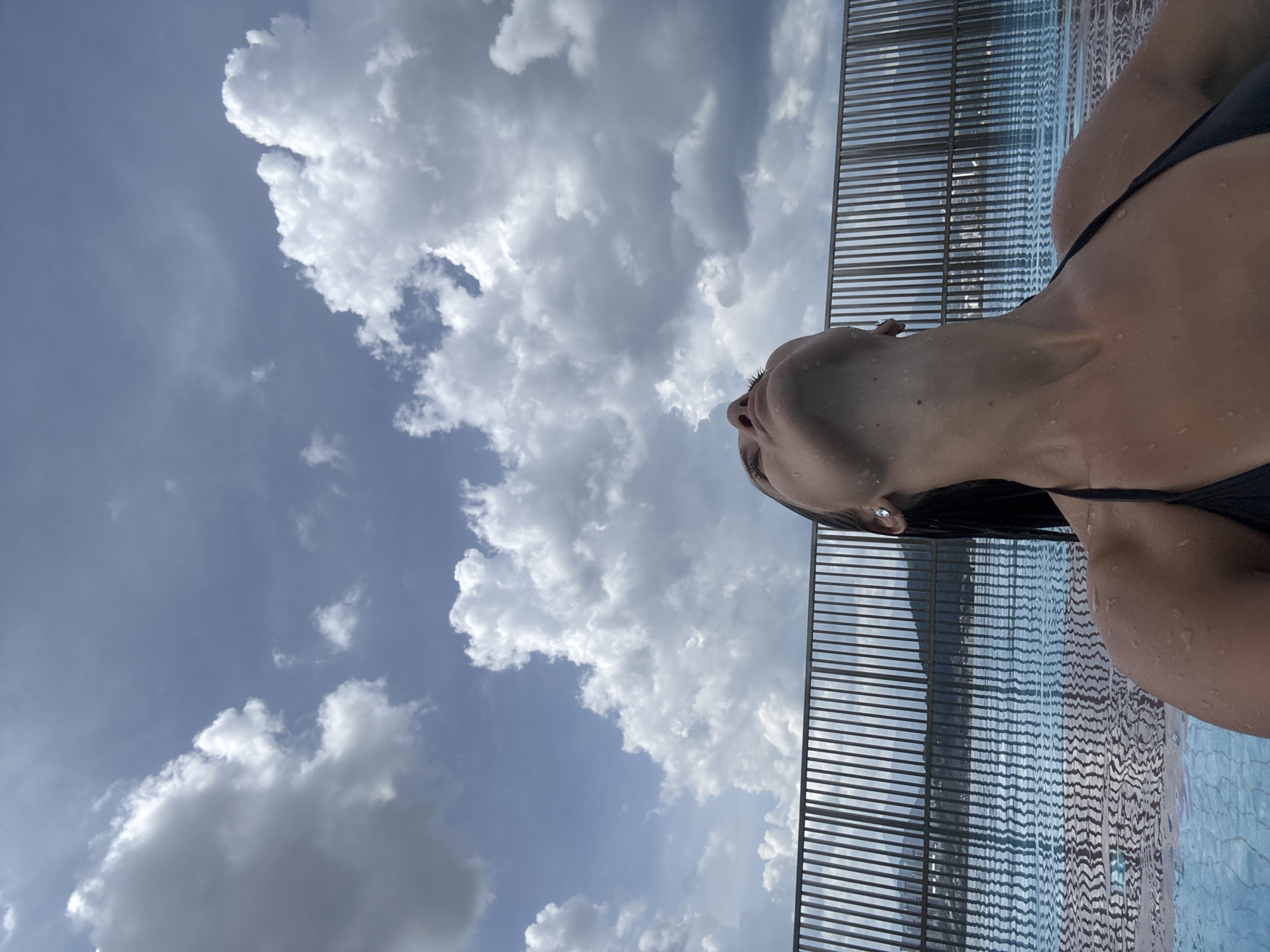 The image captures a close-up, low-angle view of a woman relaxing in a swimming pool, oriented vertically with the sky dominating the left side. The woman's upper torso, neck, and face, from below the nose, are visible on the right side of the frame. She wears a dark swimsuit, and her skin is visibly wet with water droplets. Her head is tilted back, with her gaze directed upwards towards the sky.
The setting is an outdoor swimming pool, with the water's surface, showing ripples and reflections, visible alongside the woman. A modern, dark-colored slatted fence or railing runs horizontally across the top-right portion of the image, bordering the pool area. The background is a bright blue sky filled with numerous large, fluffy white and grey cumulus clouds, indicating a partly cloudy day. The bright lighting suggests it is daytime. The overall impression is one of relaxation and contemplation. No text is visible in the image. The location is Ampang Jaya Municipal Council, Malaysia.