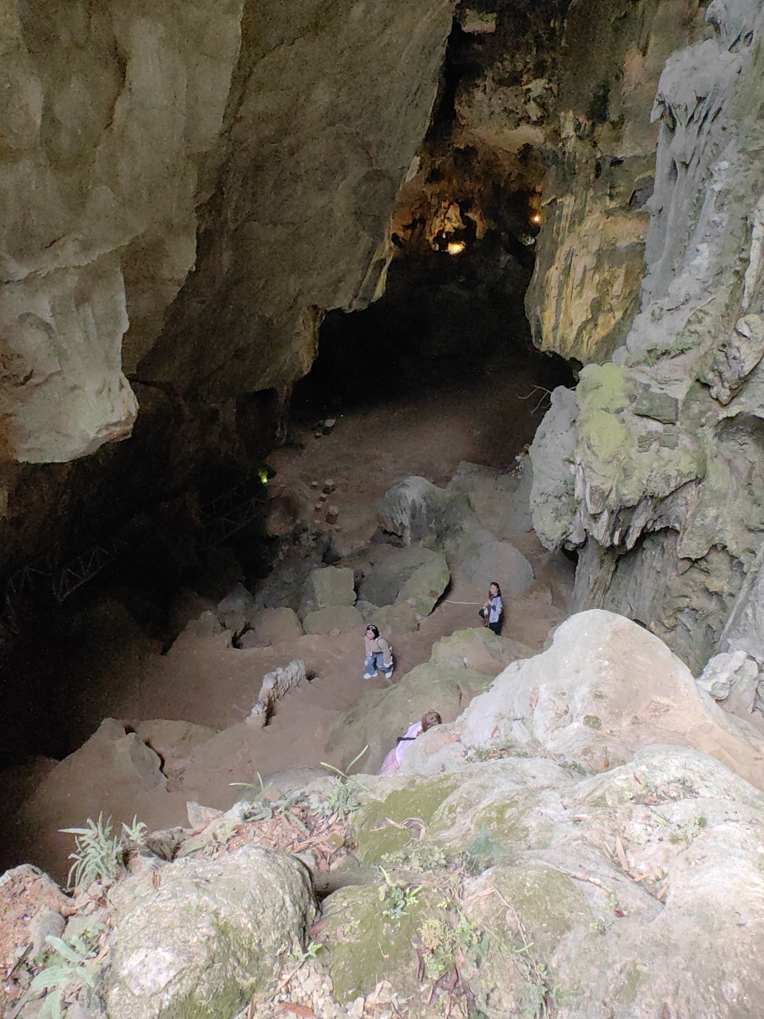 The image shows the entrance to a cave in Lạng Sơn, Vietnam. The cave entrance is framed by large, gray rock formations, some covered with green moss. Inside the cave, the terrain descends steeply, with a dirt path and scattered rocks. Three people are visible on the path within the cave. In the background, faint light suggests a deeper part of the cave.